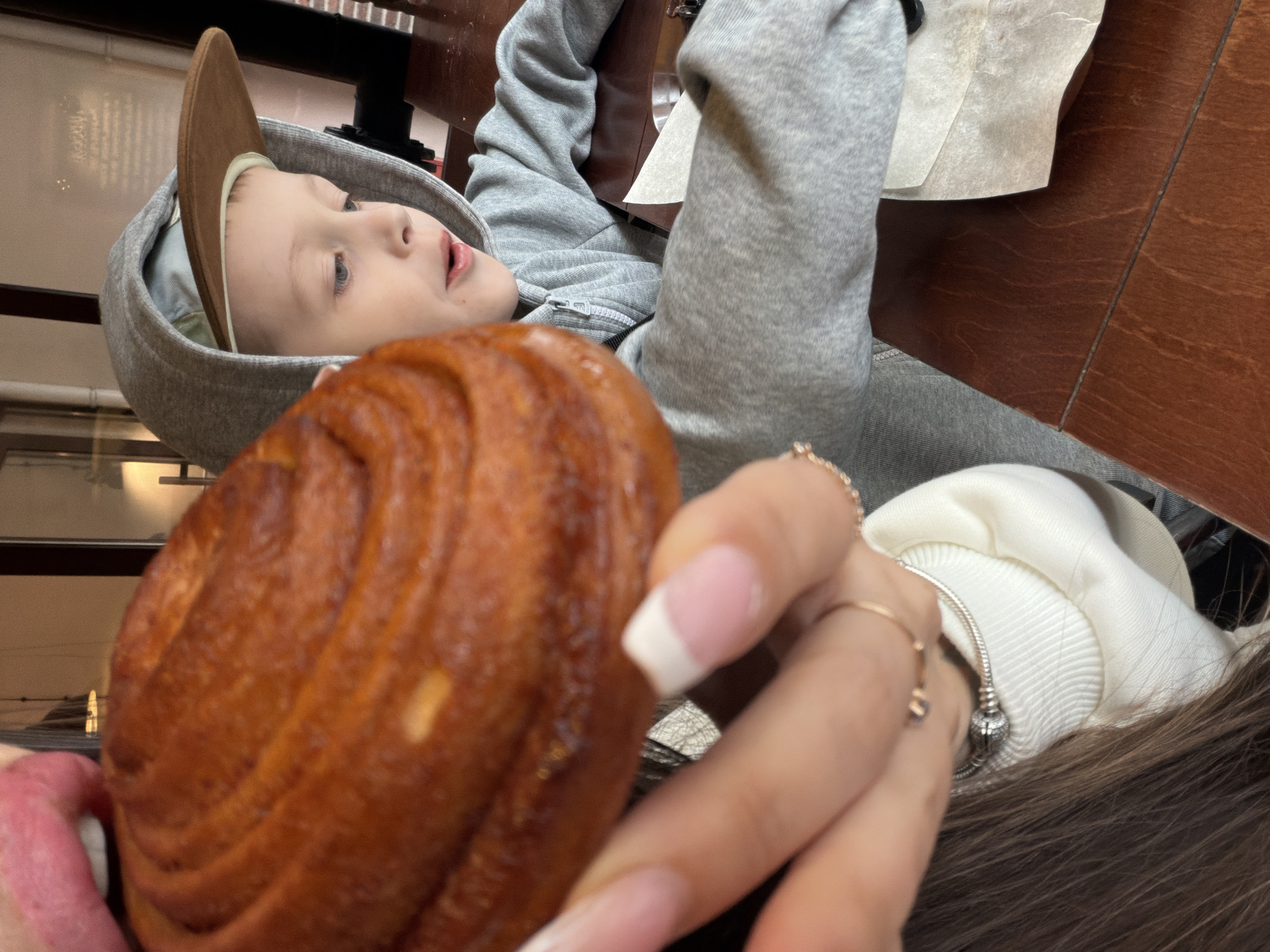 The image shows a child in a gray hooded sweatshirt and brown baseball cap looking upward, with a large cinnamon roll in the foreground near a partially visible mouth. A hand with manicured nails and rings holds the pastry. The setting appears to be a table with a wooden surface. Due to the absence of identifying landmarks, a specific location in Saint Petersburg, Russia, cannot be determined from the image.