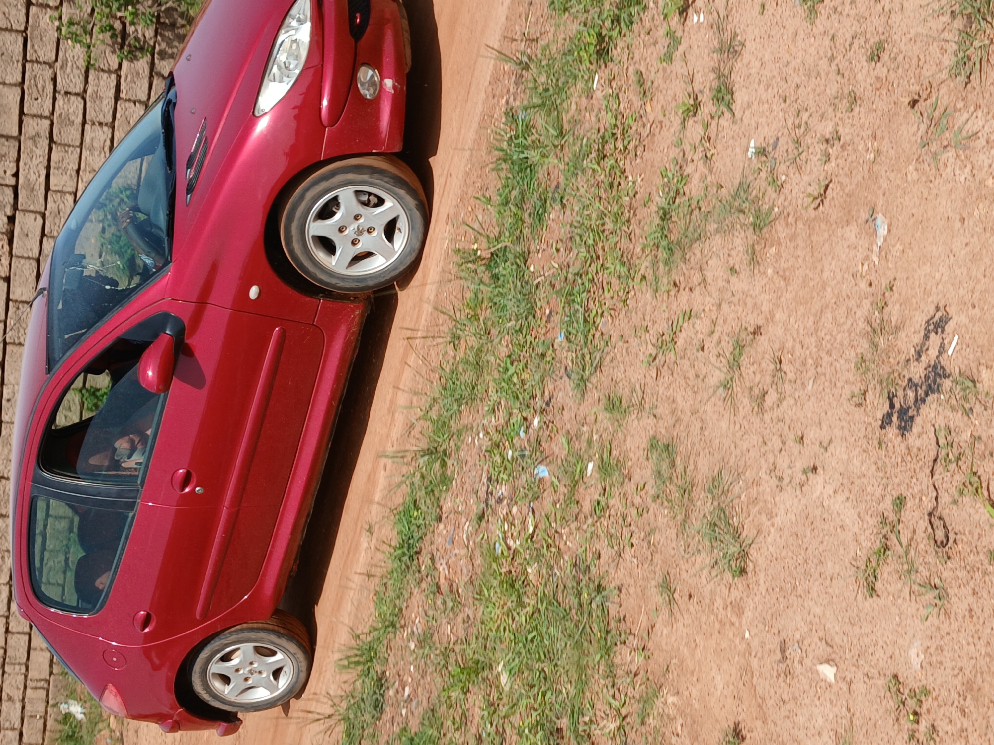 A red Peugeot 206 is parked on a dirt road in Kwang, Nigeria. The car is facing to the right of the frame and is slightly angled towards the viewer. The driver's side door is open and a person can be seen sitting in the driver's seat. The car is clean and appears to be in good condition. The road is made of dirt and is somewhat uneven. There is some grass growing on the side of the road. In the background, there is a brick wall. The sky is not visible in the image.