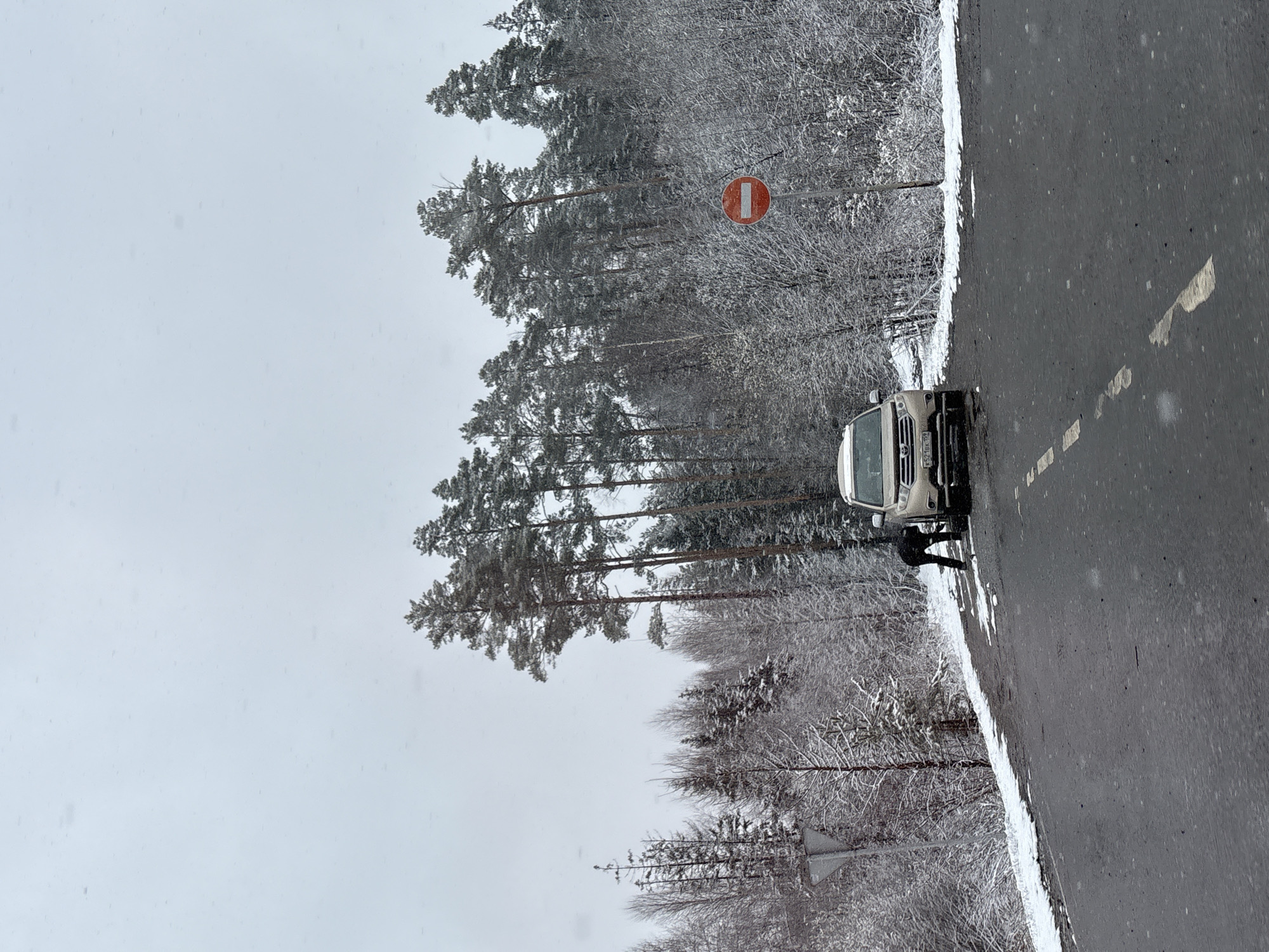 The image depicts a winter scene near Krasnokamsk, Russia. A white vehicle is parked on the edge of an asphalt road. Next to the car, a person is discernible, though their actions are unclear. Snow covers the ground, particularly along the road's edge and within the adjacent forest. A "No Entry" road sign is visible on a post near the trees. The sky is overcast, suggesting snowy weather.