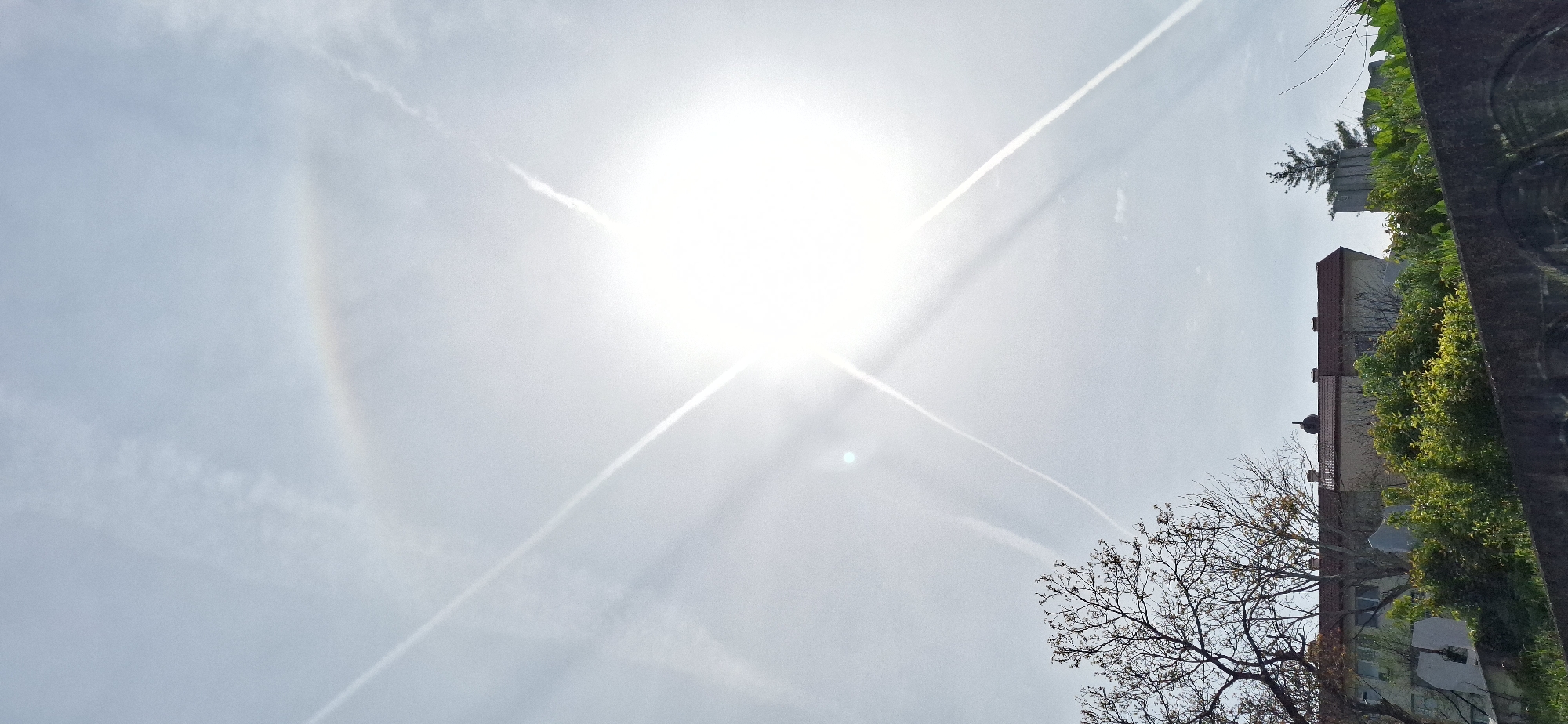 The image depicts a bright, overcast sky dominated by the sun, appearing as a large, white disc. Several contrails from airplanes intersect the sky, forming a crisscross pattern around the sun. A faint rainbow-like arc is visible to the left of the sun.
On the right side of the image, there are partial views of buildings and trees, which are rotated sideways. The buildings are brick with visible windows. The trees have both green foliage and bare branches, suggesting it may be autumn or early spring.
Without more context, it's difficult to determine the exact location within Blagoevgrad. Further analysis of building styles or prominent landmarks would be needed to pinpoint the specific area.