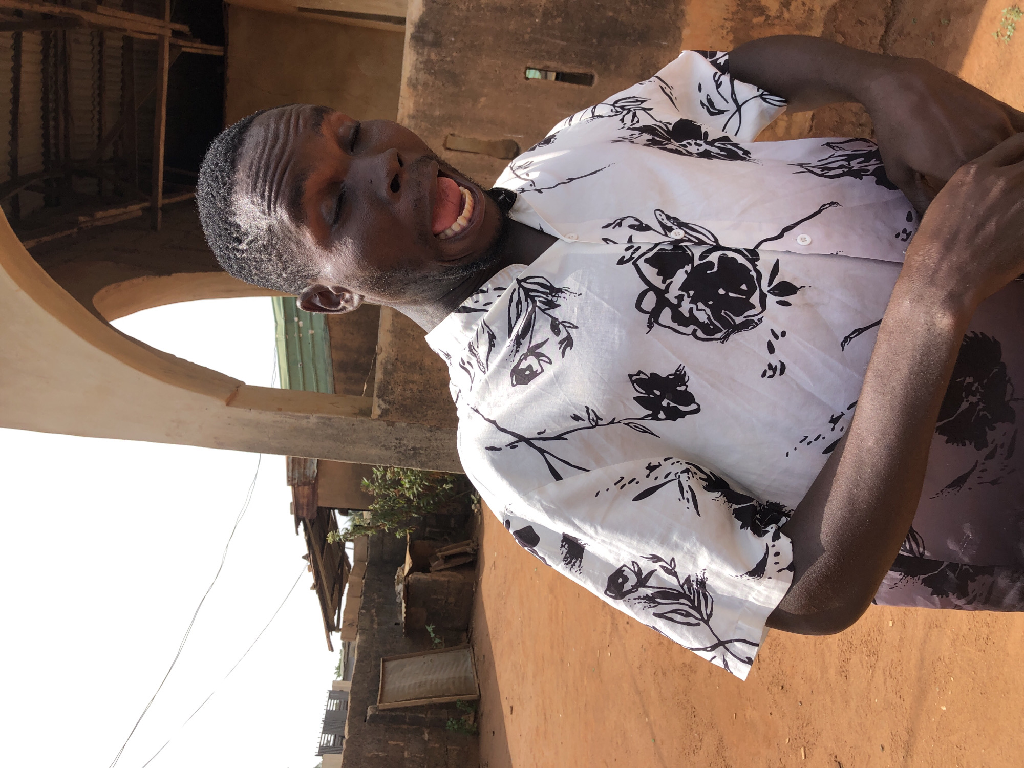 A medium shot depicts a dark-skinned man with short, cropped hair wearing a white short-sleeved shirt adorned with black floral patterns. The man's eyes are closed, his mouth is open, and his expression suggests either laughter or a yell. He is standing outside in an area with buildings that appear to be in a state of disrepair. The ground is reddish-brown, suggesting a dirt or clay surface. The photograph appears to be taken during the day with ample sunlight. The city and specific location within the city are unknown.