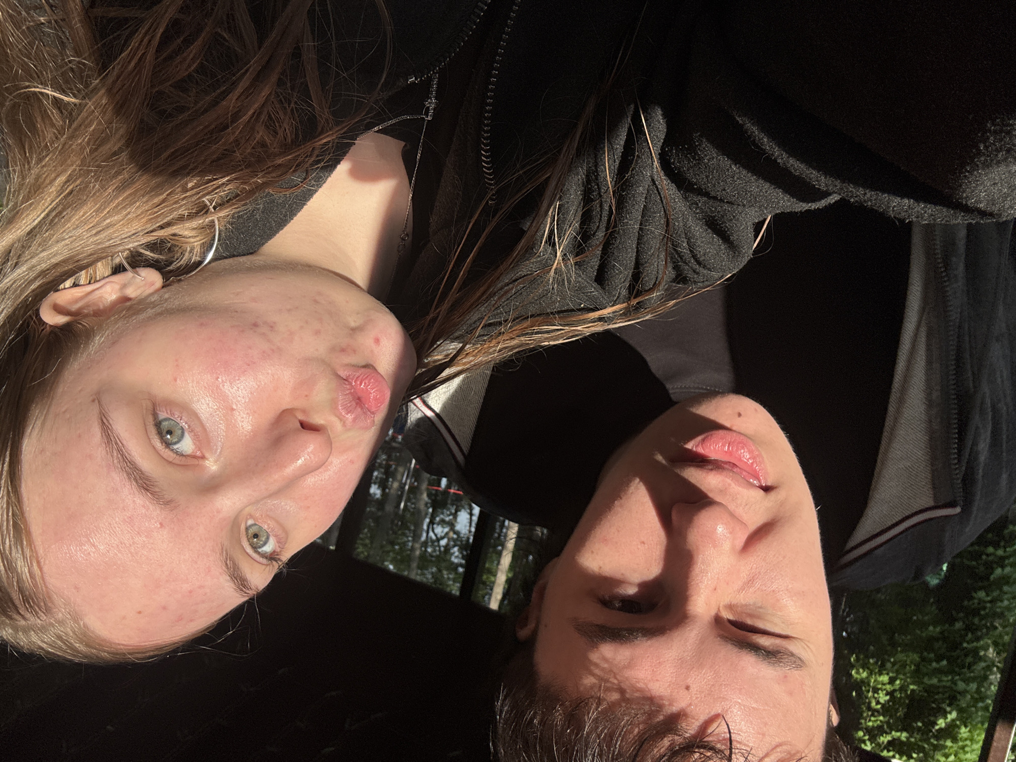 Two people are in the image. The person on the left has light brown hair, fair skin, and blue eyes. They are wearing a black hooded jacket, and a silver earring is visible in their left ear. They are pursing their lips slightly. The person on the right has short dark hair, fair skin, and an eye that is partially closed. They are also wearing a black jacket over a grey shirt with a white collar. Trees and foliage are visible in the background, partially obscured by what appears to be a car window. The picture has strong lighting that casts shadows across the faces. The exact location within the city of Perm is not discernible from the image.