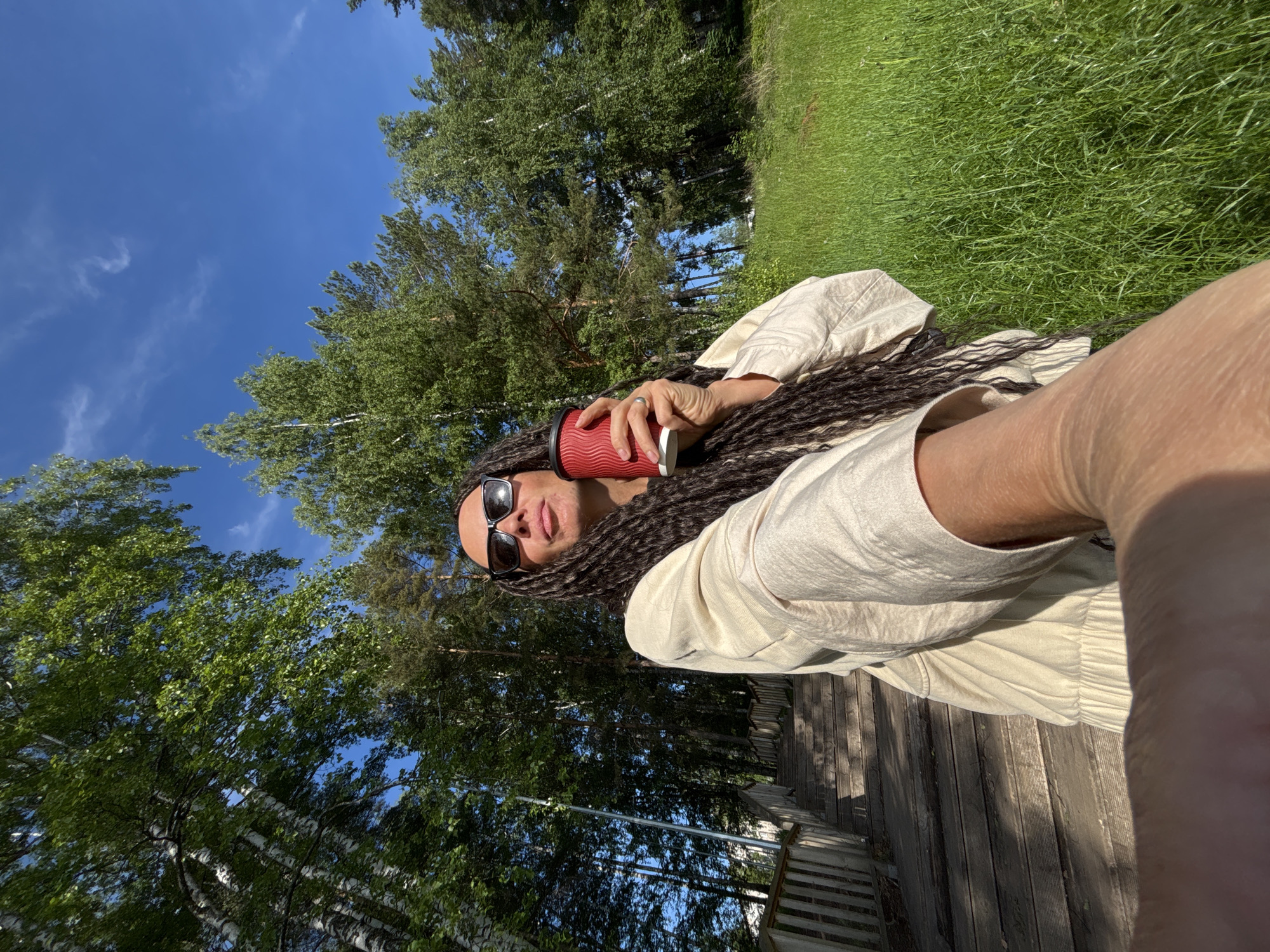 A person is captured in a selfie outdoors on a sunny day, likely in Chaikovsky, Russia. The individual has long, dark dreadlocks or braids and is wearing sunglasses, which obscure part of their face. They are dressed in a light-colored, possibly off-white, long-sleeved shirt. In their hand, they hold a textured red reusable coffee cup with a white lid.
The background reveals a vibrant natural setting. Above the person, a clear blue sky with a few thin, wispy clouds is visible. Lush green trees, including some identifiable birch trees with their distinct white bark and other conifers, surround the area, suggesting a forest, park, or a spacious garden. To the side, a patch of bright green grass is visible, and beneath the person, there's a wooden deck or pathway, hinting at an outdoor seating area or structure. The bright sunlight casts distinct shadows, indicating a clear and pleasant weather day. The overall scene conveys a sense of relaxation and enjoyment of the outdoors. No visible text is present in the image.