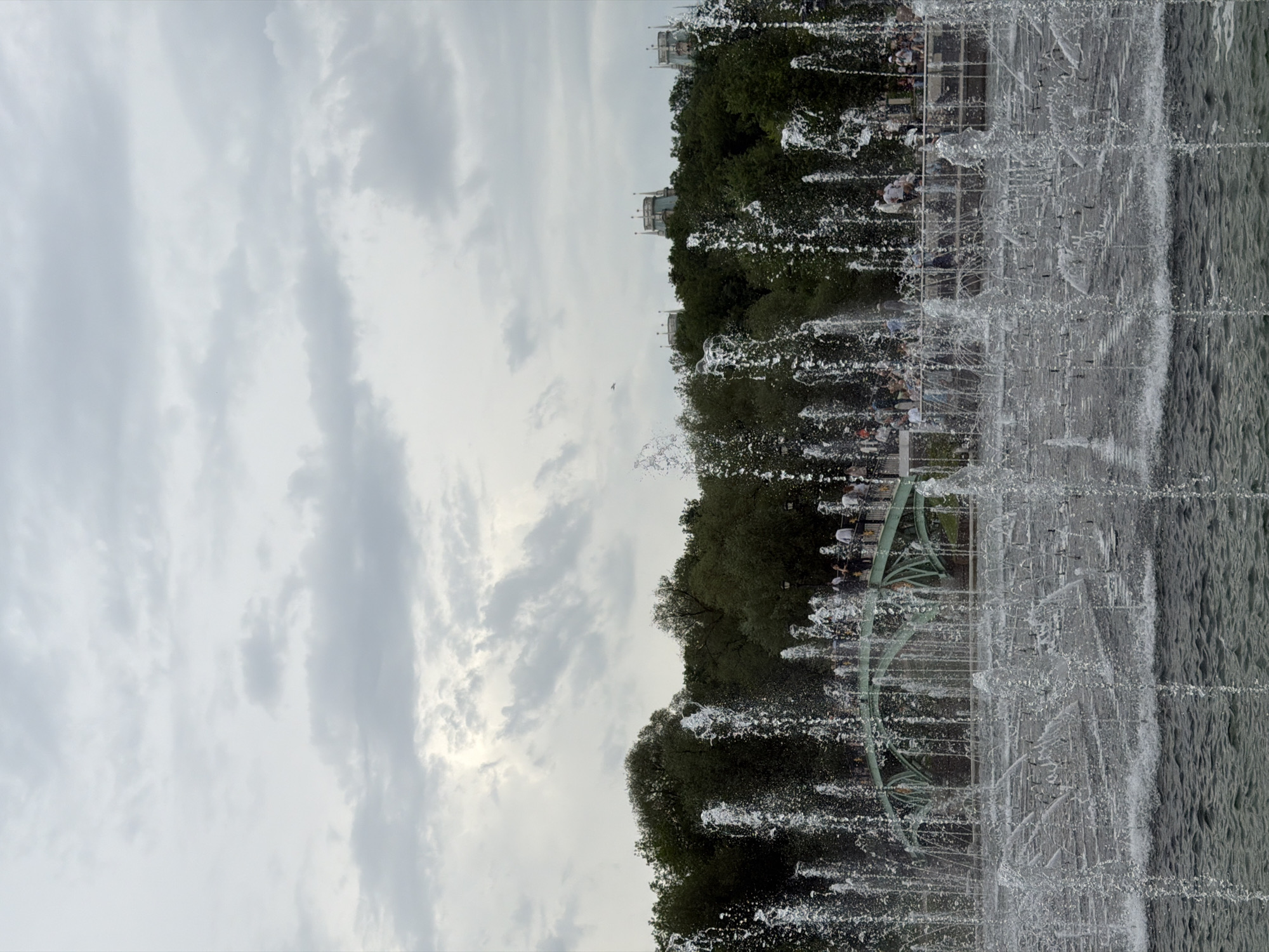 The image shows a fountain in Moscow, Russia, viewed from a ground-level perspective. The fountain is actively spraying water, obscuring some of the background. Behind the fountain, trees and buildings are visible. People can be seen walking near the fountain. The sky is overcast with grey clouds.