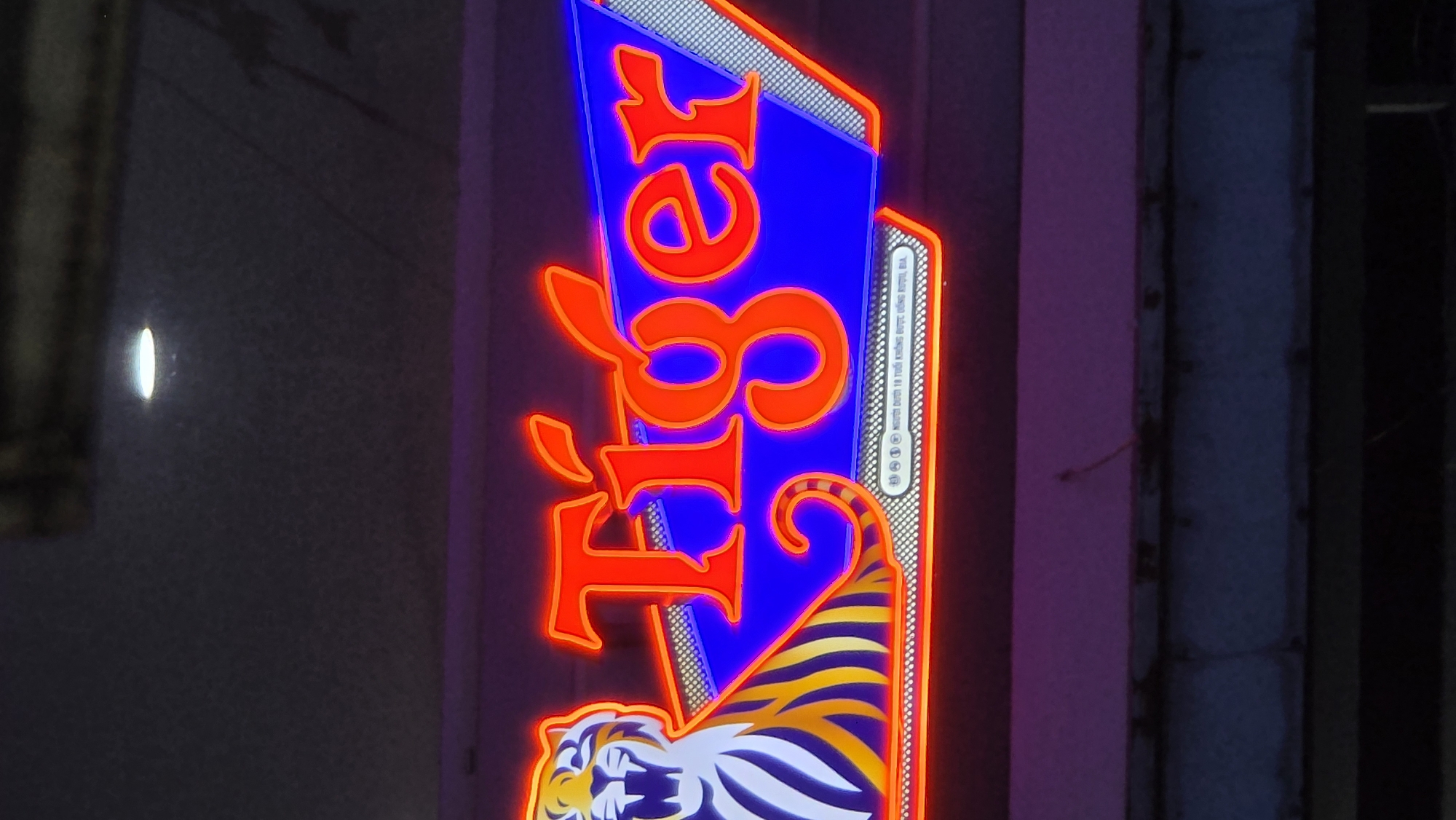 A close-up shot depicts a vertical Tiger Beer sign in Nha Trang, Vietnam. The word "Tiger" is displayed in red neon against a blue background, with a tiger illustration at the bottom of the sign. The sign is positioned against a dark backdrop with glimpses of surrounding structures.