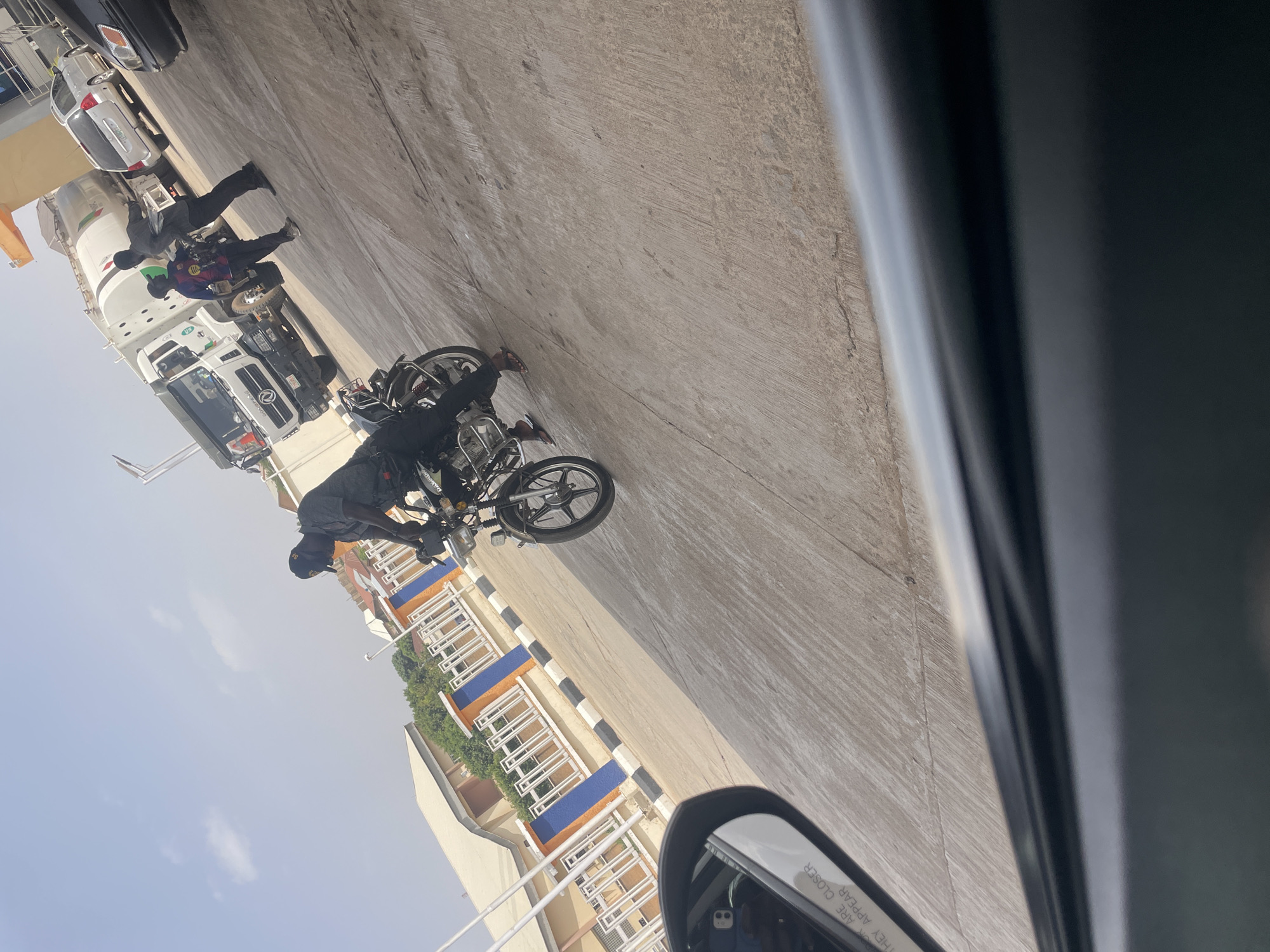 The image depicts a street scene in Jos, Nigeria. A motorcycle is parked on the side of the road. Two men are standing near a white truck. A building with white balconies and orange and blue accents is visible in the background. Cars are parked further down the street. The photo was taken from inside a car, with part of the car door and side mirror visible in the foreground.