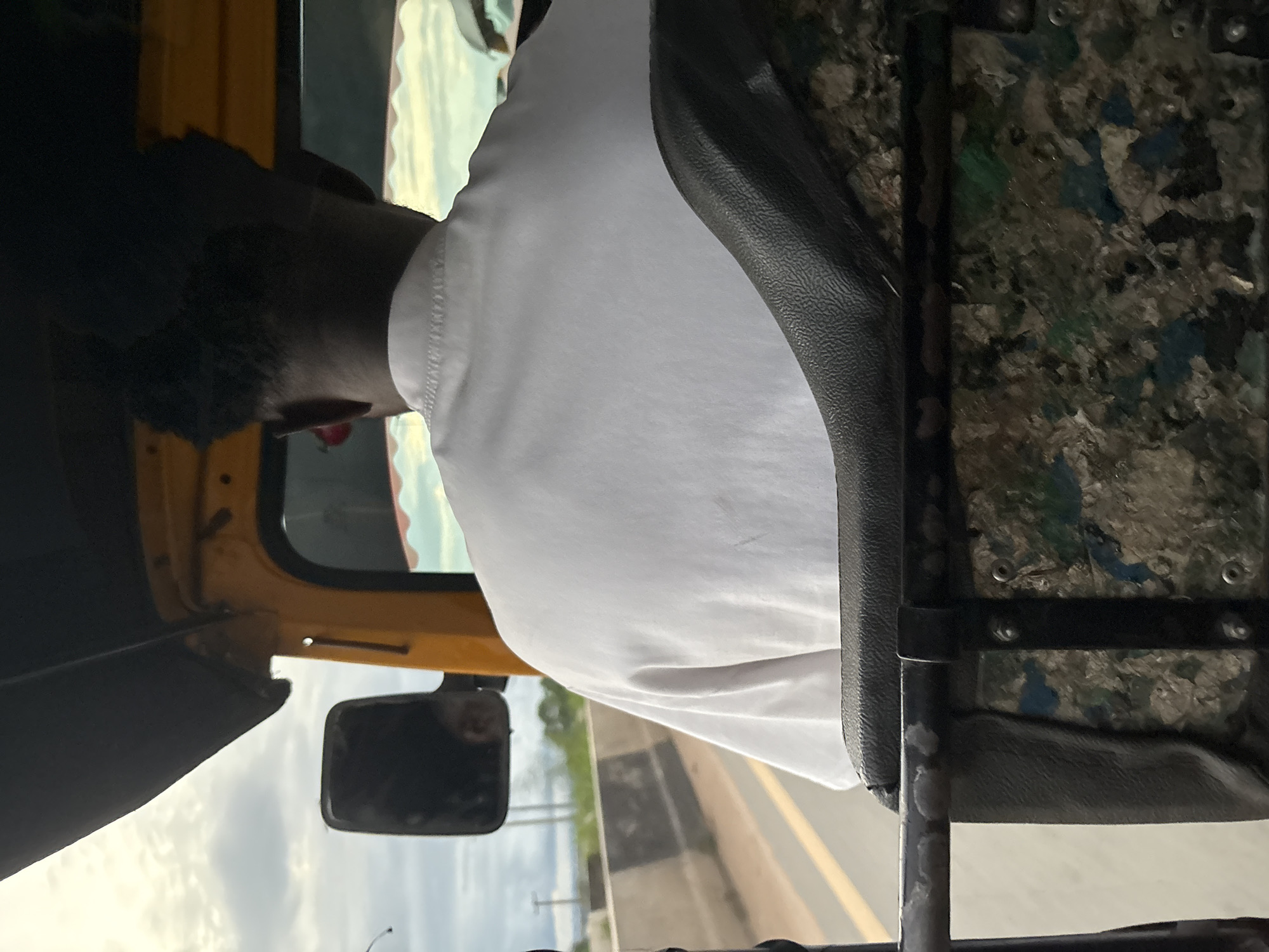 The image shows the interior of a yellow auto-rickshaw, a common mode of transport in Onitsha, Nigeria. The camera is positioned behind the driver, capturing the back of his head and shoulders. He is wearing a white short-sleeved shirt and has dark, curly hair. The auto-rickshaw's interior is visible, including a black upholstered seat and a metal bar with a patterned backing. Through the open sides of the vehicle, a road with a concrete barrier and trees can be seen, indicating movement and a roadside environment. The sky above is cloudy.