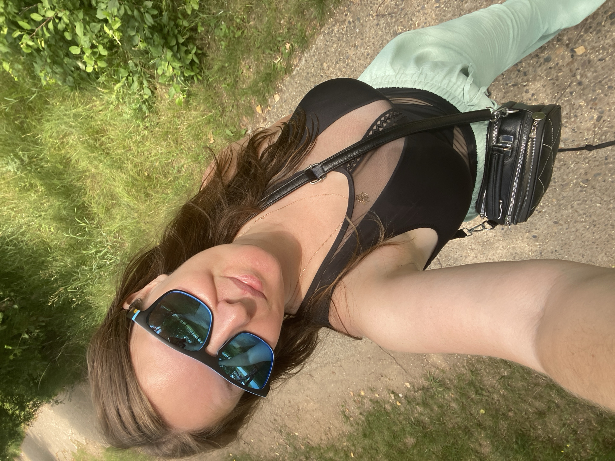 In a selfie taken outdoors on a path lined with grass and trees, a woman with brown hair wears sunglasses, a black top with sheer mesh detailing, light green pants, and a black crossbody bag. The scene is set in Noviy, Russia, under natural lighting.