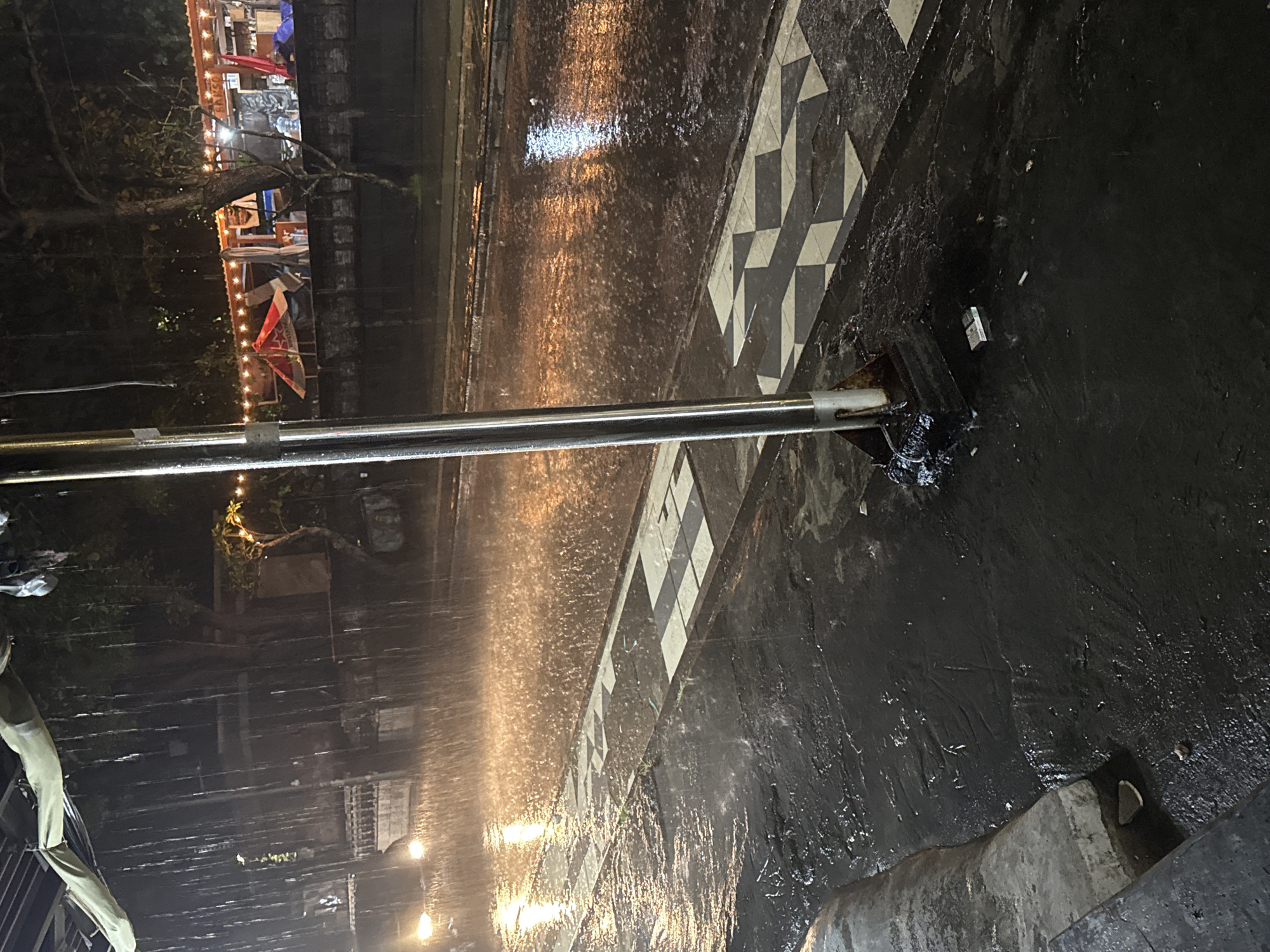 This media file depicts a rainy night scene in Kuta, Indonesia, viewed from a slightly elevated perspective. The main subject is a wet, dark street or pathway, glistening with reflections from artificial lights. A prominent feature is a section of pavement adorned with a distinctive black and white geometric pattern, resembling tessellated squares or diamonds, which appears to be a crossing or walkway. A metallic railing or bar extends horizontally across the middle ground, separating the viewer from the street level.
It is actively raining, with visible streaks of water and splashes on the glistening surfaces. Small pieces of litter, including what appears to be cigarette butts and a small rectangular object, are scattered on the wet ground near the railing's base. In the left background, an establishment, possibly a cafe or restaurant, is visible, illuminated by warm string lights strung along its exterior. Dark trees and foliage surround this building. Faintly visible on a sign above the lights is the word "BAKSO," suggesting the establishment serves Indonesian meatballs. The time of day is night or late evening, as indicated by the reliance on artificial lighting and the dark surroundings. The scene evokes a moody, atmospheric ambiance typical of a rainy urban night. No people are visible in the image.