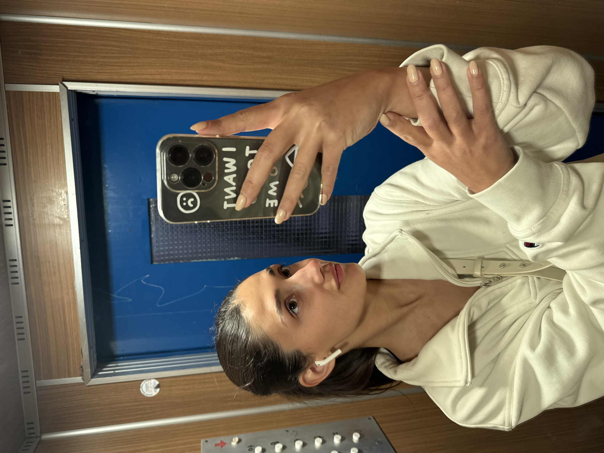 A young woman is captured taking a mirror selfie, likely within an elevator or a similar enclosed space. She has dark hair pulled back, dark eyes, and a neutral, contemplative expression. She is wearing a cream or off-white zippered hooded sweatshirt and has a white AirPod in her right ear. Her nails are manicured with a light, nude polish.
She holds a smartphone, identifiable by its triple-lens camera system, in her left hand, pointed towards her reflection. The phone case is clear or translucent with black accents, displaying a reflected and inverted message that reads "I WANT TO GO HOME," accompanied by a stylized smiley face with a question mark for its mouth. Her right hand is positioned across her left forearm.
The setting features walls with horizontal wood-grain paneling and metallic vertical and horizontal trim. Reflected in the mirror behind her is a deep blue surface, possibly a door or wall, showing some lighter, wavy marks. At the bottom of the frame, a metallic elevator control panel is visible with multiple white circular buttons, one partially showing the number "10," and a red arrow indicator. The lighting is artificial, suggesting an indoor environment. The scene takes place in Bar, Montenegro.