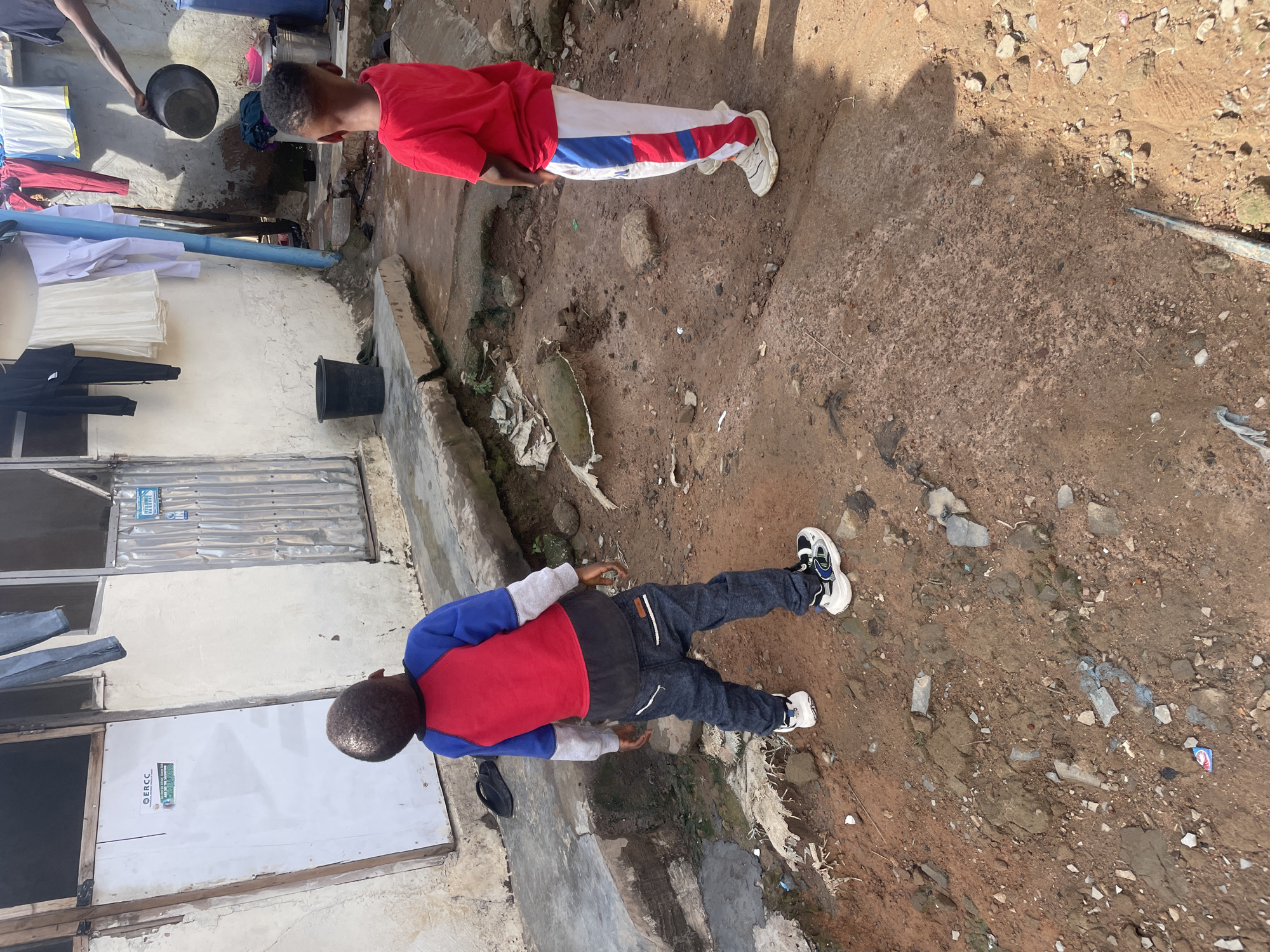 Two young boys stand in an outdoor area with a rough, dirt and concrete ground, possibly in a residential compound in Abuja, Nigeria. The boy on the left, facing away from the camera, wears a blue and red long-sleeved shirt and dark denim jeans. He is looking upwards and to the left. The boy on the right, also facing away from the camera, wears a bright red short-sleeved shirt and white pants with red and blue stripes down the sides. He is looking towards the left. In the background, there is a light-colored building with corrugated metal elements and a white door with signage. Laundry hangs on a line above and to the right of the boys. Various objects, including buckets and debris, are scattered around the ground. The scene appears to be a casual, everyday setting.
