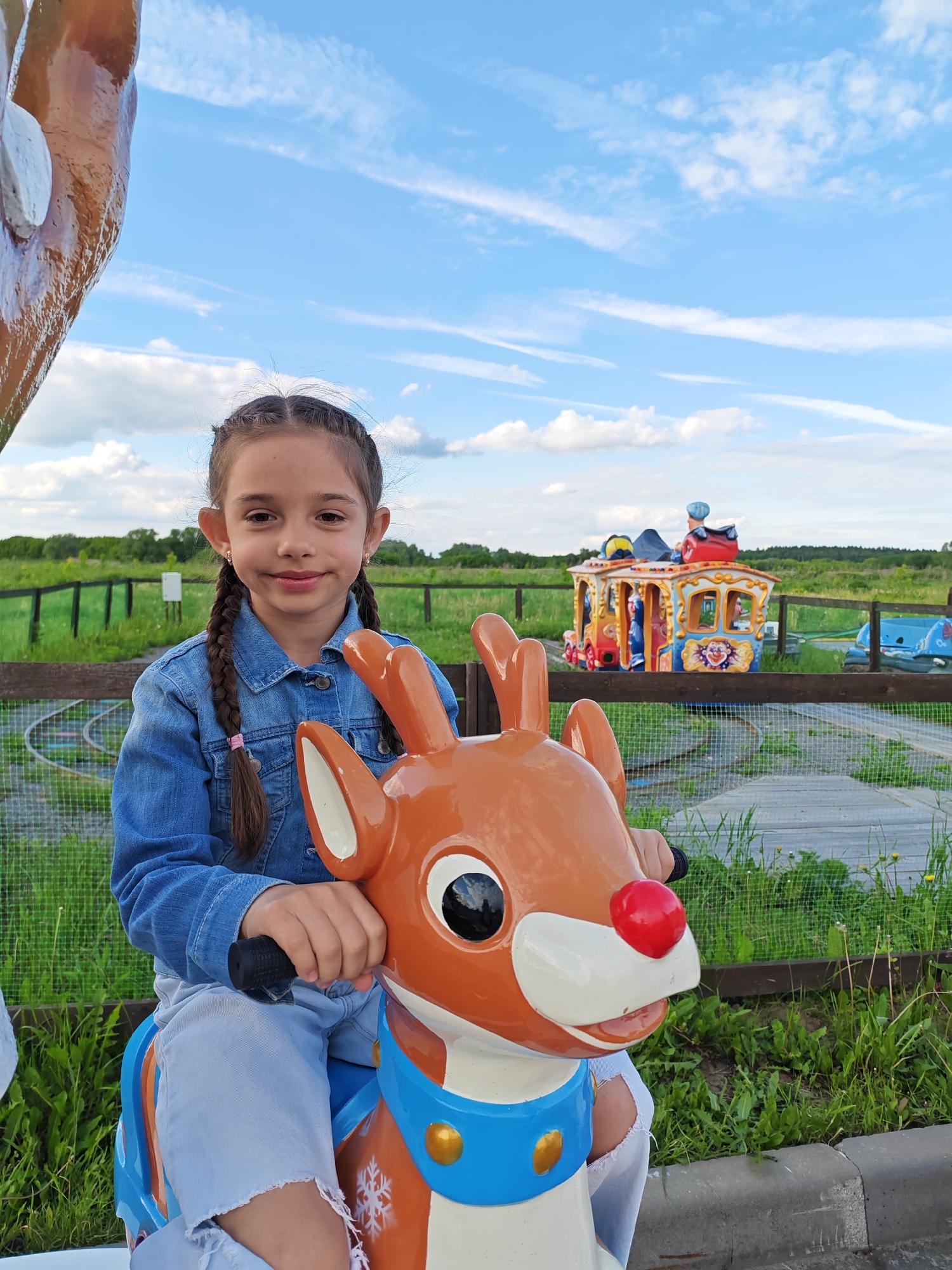 In Bronnitsy, Russia, a young girl with braided brown hair and a denim jacket sits on a coin-operated ride shaped like a reindeer with a red nose. The reindeer is brown and white with a blue collar. The setting is an outdoor amusement area with grass, fences, and a colorful train ride in the background. The sky is blue with scattered clouds.