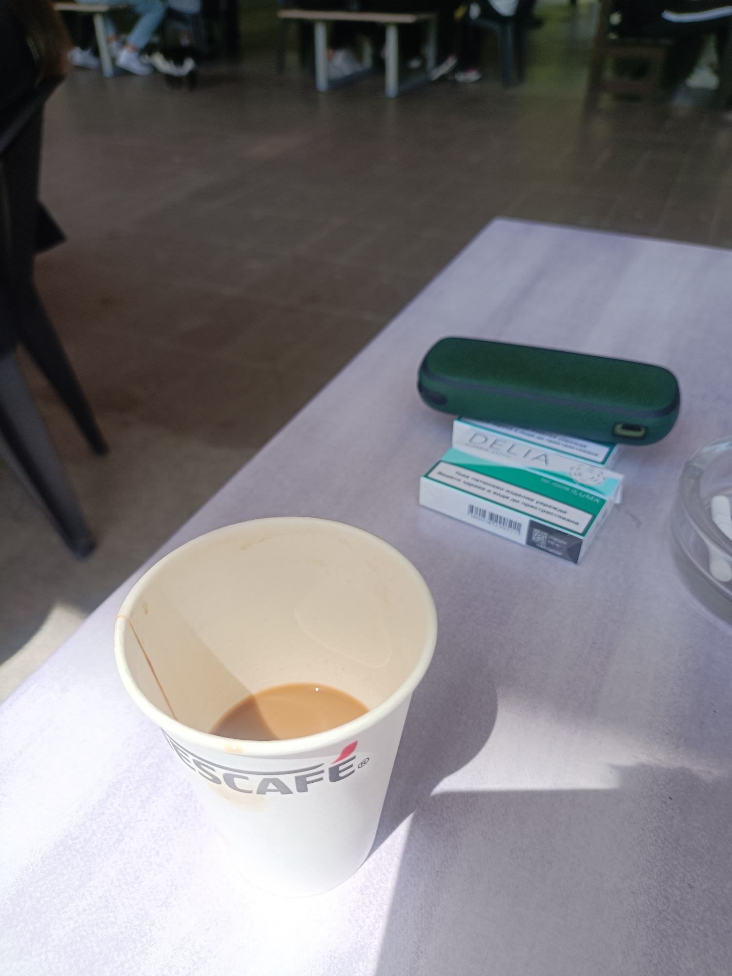 The image presents a close-up, high-angle view of a table in an indoor or semi-outdoor public setting, likely a cafe or common area, in Plovdiv, Bulgaria. The scene is brightly lit by natural light, casting clear shadows on the table, indicating it is daytime.
In the foreground, a white paper cup with the red "NESCAFE®" logo is partially filled with a light brown beverage, suggesting coffee or tea with milk. Beside the cup, on a light-colored table surface, are items associated with heated tobacco use. A dark green, rectangular protective case, possibly for an e-cigarette or heated tobacco device, rests atop two white boxes. The boxes are labeled "DELIA" and feature Cyrillic text: "Това тютюнево изделие уврежда Вашето здраве и води до пристрастяване." (This tobacco product harms your health and leads to addiction.), alongside "for IQOS ILUMA" and a barcode, identifying them as tobacco sticks. To the right, a clear ashtray holds several white cylindrical sticks, consistent with heated tobacco residue.
The background is out of focus, showing blurry figures of people, with legs and feet (some wearing white sneakers) visible under tables and chairs, seated in what appears to be a casual gathering space. The floor is a dark, possibly concrete or tiled surface. The overall impression is of a relaxed, informal setting where people are taking a break and socializing.