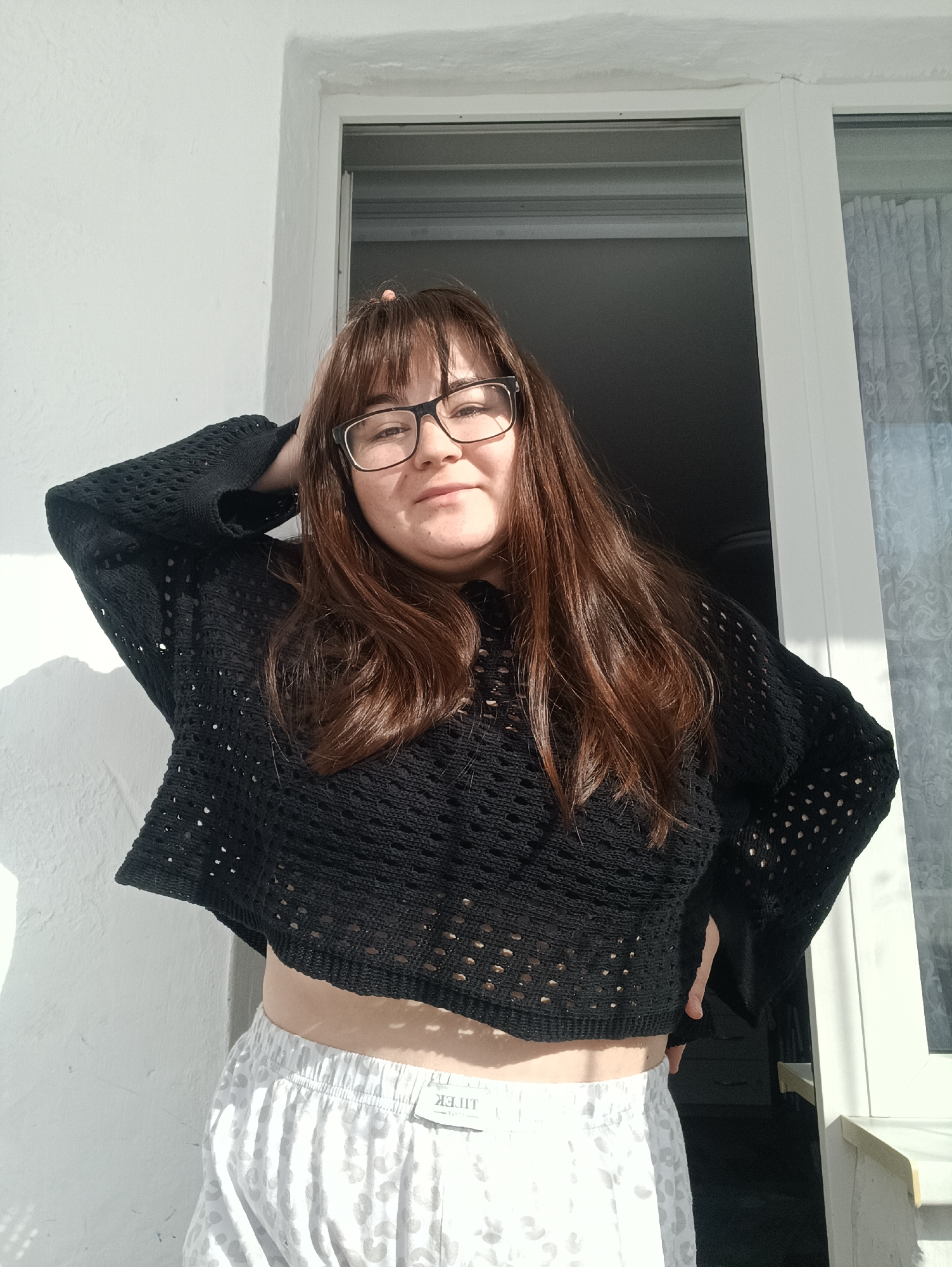 A female subject with medium-length brown hair and bangs, wearing rectangular black-framed glasses, stands partially illuminated by strong directional sunlight. She has a light complexion and a slight smile, looking directly at the viewer. Her right arm is raised with the hand placed behind her head, and her left hand rests on her hip. She is dressed in a black, long-sleeved, open-knit (crocheted) crop top that exposes her midriff, paired with light-colored shorts featuring a grey/white leopard print pattern and an elastic waistband. A clothing tag, possibly displaying "LITIEK" in reverse, is visible on the shorts. The background consists of a textured, off-white wall on the left, transitioning to a white PVC window or door frame in the center and right. Beyond the frame, a dark interior space is visible, with a sheer, light-colored patterned curtain partially visible on the far right. The specified location of Noviy, Russia, cannot be confirmed from the visual evidence within the image.