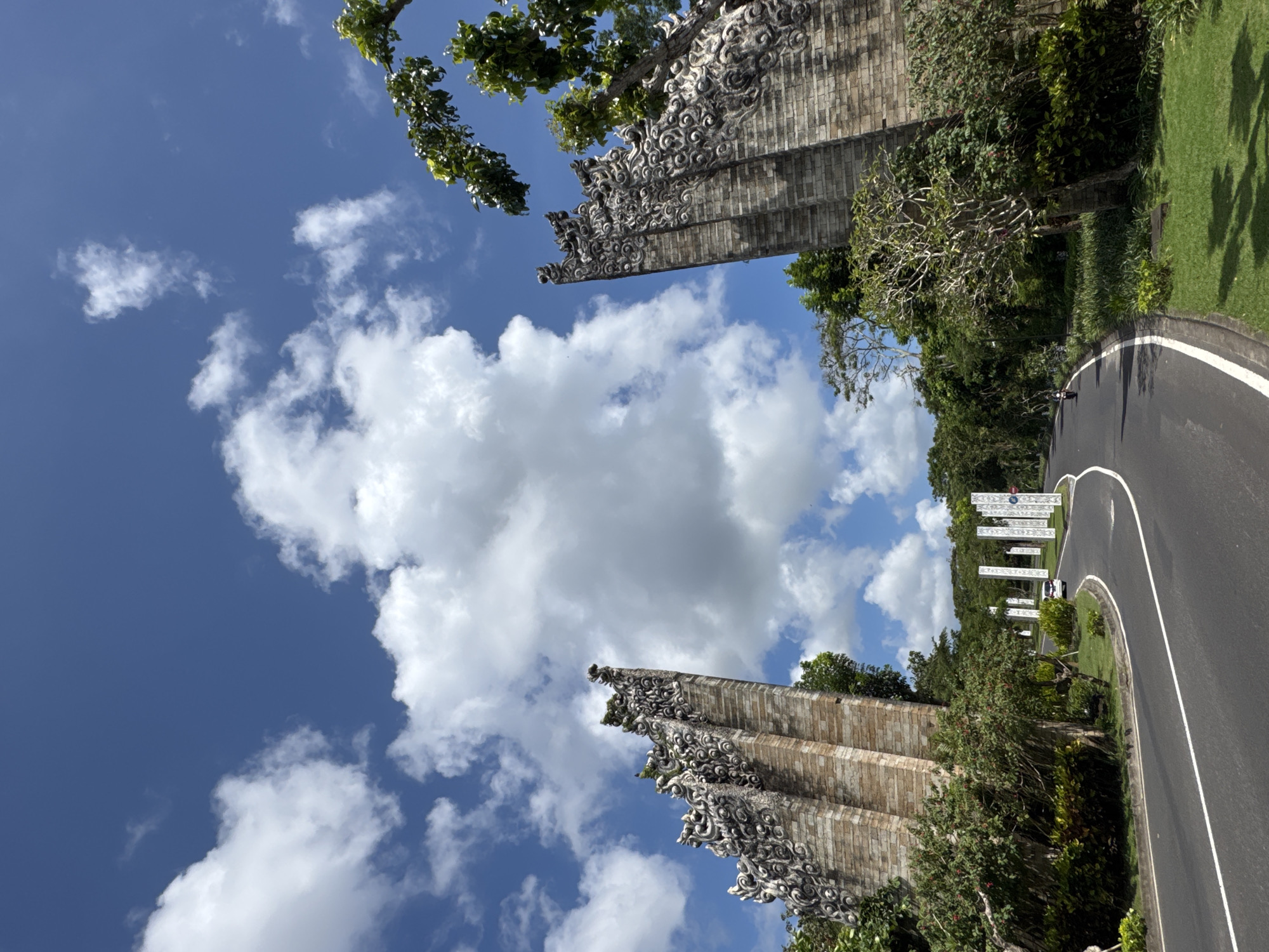 This outdoor, eye-level shot, taken in Ungasan, Indonesia, presents a vibrant scene under a bright blue, partly cloudy sky. Two ornate, brick-like stone pillars, adorned with intricate carvings at their peaks, rise from either side of the frame, their structures contrasting with the sky. Green foliage, including trees and shrubs, fills the spaces around the pillars and along a curved, asphalt road to the right. The road is marked with a white line and curves into the distance, passing a series of white posts or signs. A small figure of a person is visible walking or standing near these posts. The landscape is lush, highlighting the natural beauty alongside the architectural elements.
