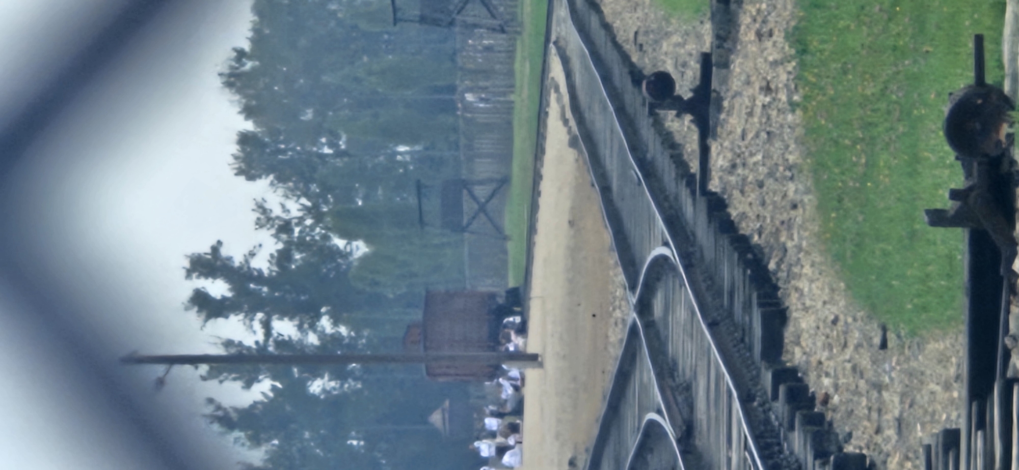 The image, taken in Brzezinka, Poland, depicts a railway track leading to a concentration camp. In the background, a wooden train car is parked on the tracks. Several watchtowers and a wooden fence are visible, suggesting a guarded perimeter. A group of people, some dressed in white shirts and dark pants, are seated or standing near the train car and along the tracks. The foreground features multiple sets of train tracks diverging and converging, with gravel and grass on either side. A switch mechanism for the tracks is visible in the lower right portion of the image. The overall scene is somber, with an overcast sky and muted colors.