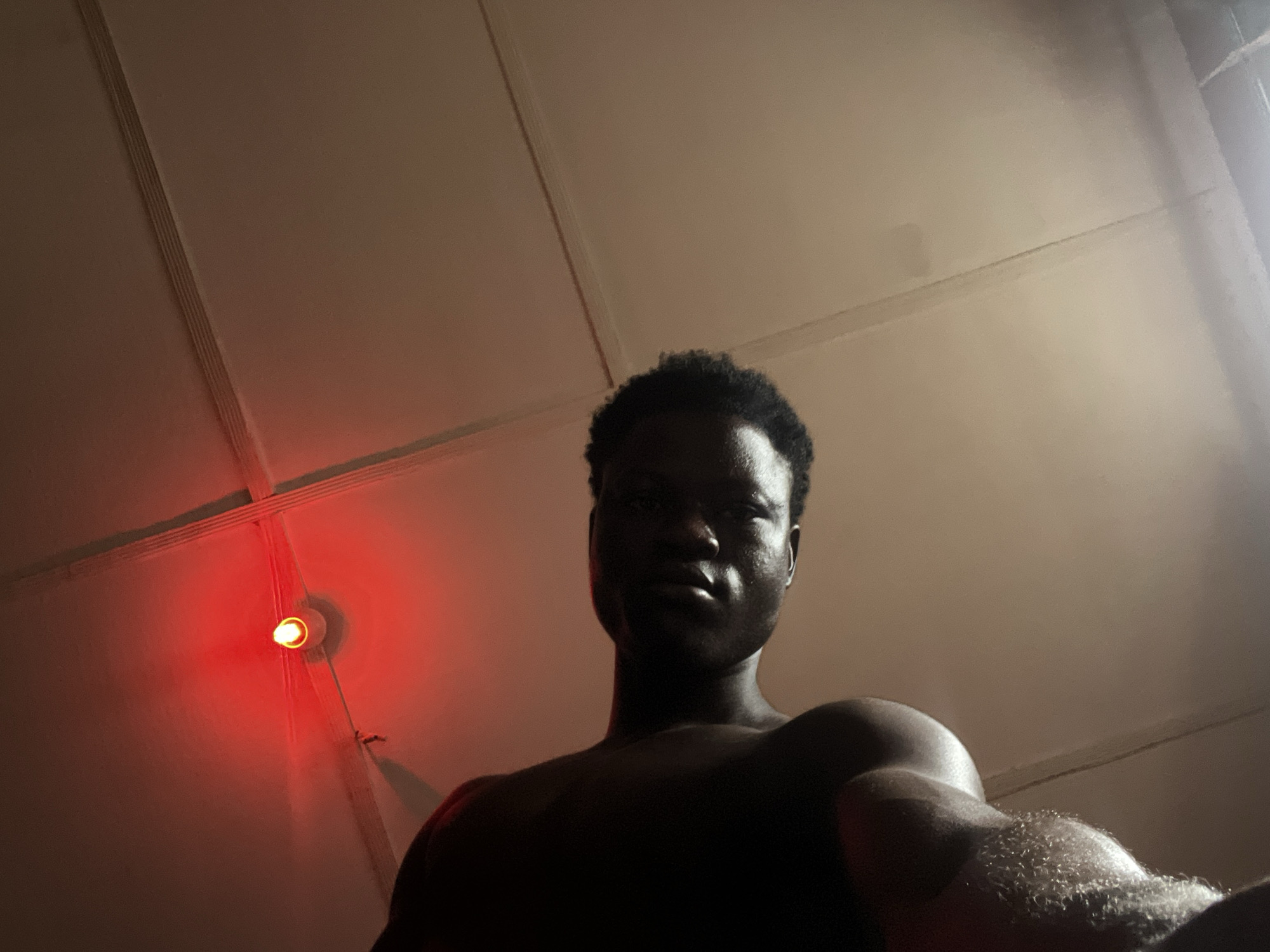 A low-angle photograph taken indoors features a shirtless Black man. The man is facing the camera. To the upper left is a ceiling and a light fixture with a red-orange bulb. The location is unknown.