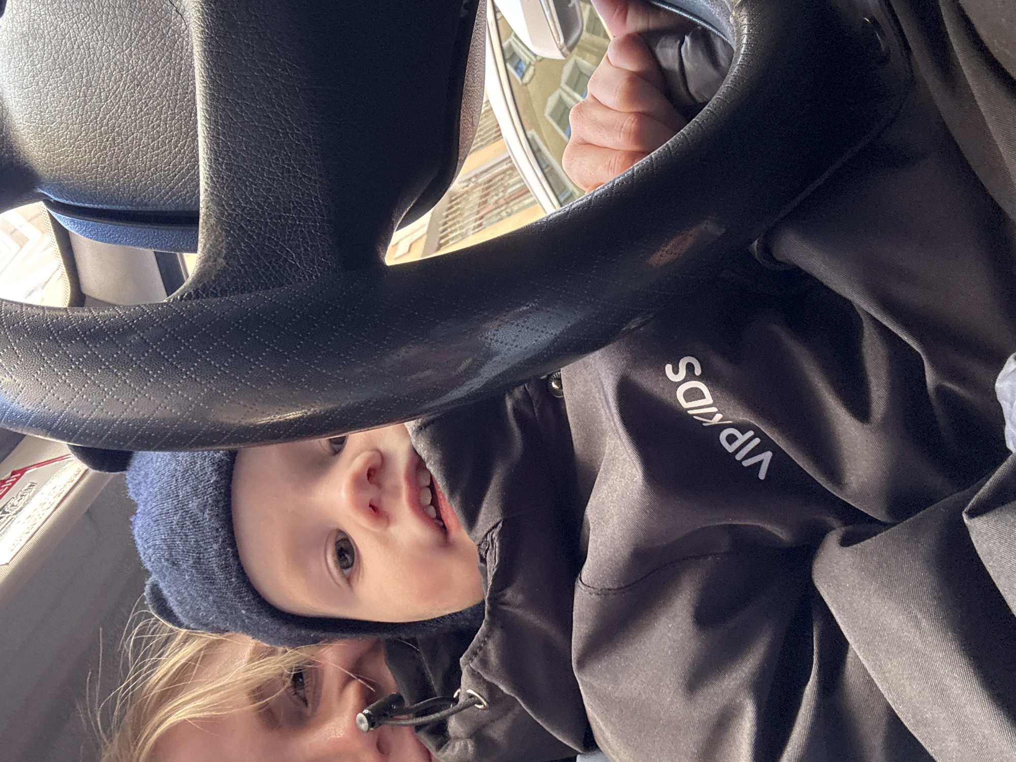 A photograph taken in Saint Petersburg, Russia, presents a close-up view from inside a vehicle. A child wearing a dark blue beanie and black jacket with the words "VIP Kids" on it peers out from behind the steering wheel. The child's face is partially visible, and they appear to be smiling. Below the child, a woman with blonde hair is partially visible. The steering wheel is dark and textured, and a hand is visible gripping it. Outside the car window, a building typical of the city's architecture can be seen.