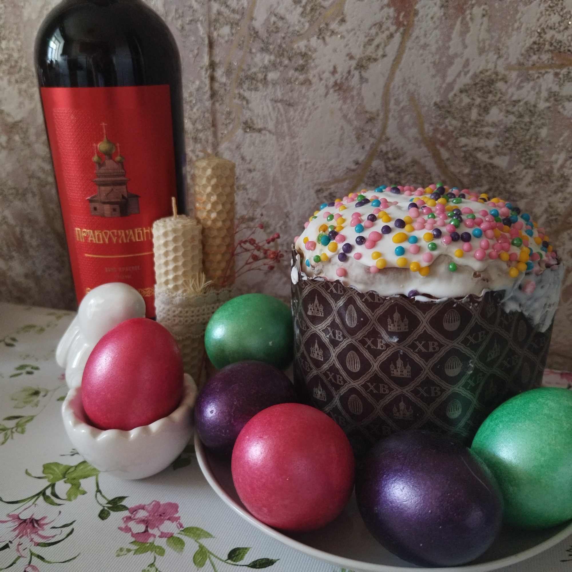 This image captures a vibrant and traditional Orthodox Easter arrangement, featuring festive elements artfully displayed on a light-colored tablecloth with a floral pattern. In the center, a large kulich (Easter bread or cake) with white icing and a generous topping of colorful sprinkles stands prominently. Its decorative paper wrapper is dark brown, patterned with the Cyrillic letters "XB" (representing "Christ is Risen"), stylized Easter eggs, and small church illustrations.
Surrounding the kulich are several brightly colored Easter eggs in shimmering shades of red, purple, and green. One red egg is nestled in a small white ceramic egg cup, next to a white ceramic bird figurine, while others are arranged on a white plate.
To the left, a dark red wine bottle is visible, featuring a red label with an illustration of a church with golden onion domes. The text on the label, in Cyrillic, reads "ПРАВОСЛАВНОЕ" (Orthodox) and "ВИНО ЦЕРКОВНОЕ" (Church Wine), indicating its ceremonial purpose. Behind the eggs and near the wine, two distinctive beeswax candles with a honeycomb pattern are present, adding to the festive and traditional atmosphere. A third candle is partially visible behind the wine bottle.
The background shows a light-colored wall with a subtle, textured, and slightly abstract pattern. The overall scene conveys a sense of celebration, tradition, and reverence associated with the Easter holiday in Orthodox Christian culture. The lighting is soft and diffused, typical of an indoor setting, and no specific time of day is discernible.