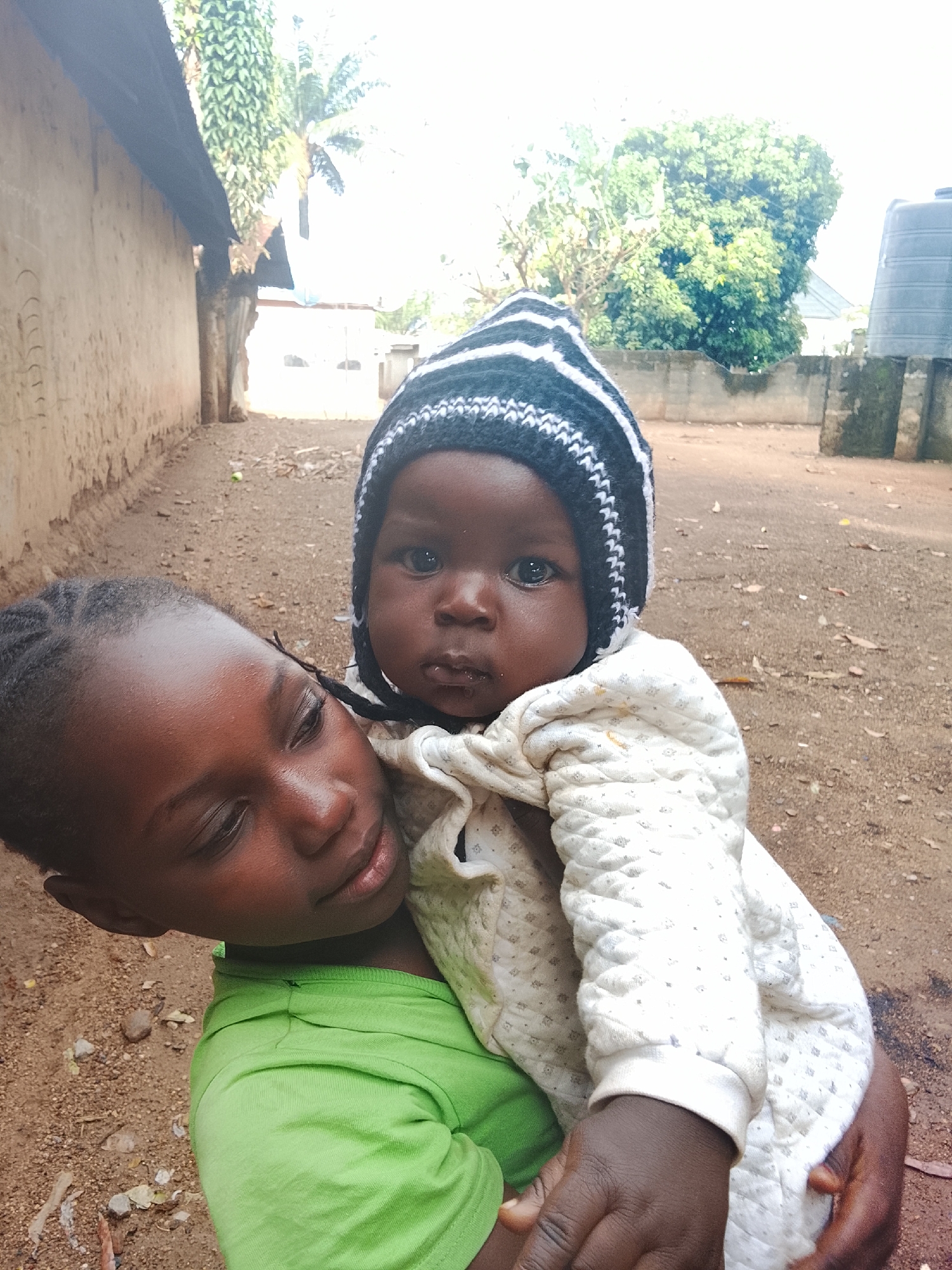 The image shows two children, one holding the other. The child being held is younger, wearing a striped knit hat and a quilted white onesie. The older child is wearing a green shirt. The setting appears to be an outdoor area with a dirt ground, a wall to the left, and trees in the background. A water tank is visible in the background. The image may be in or near Akwanga, Nigeria.