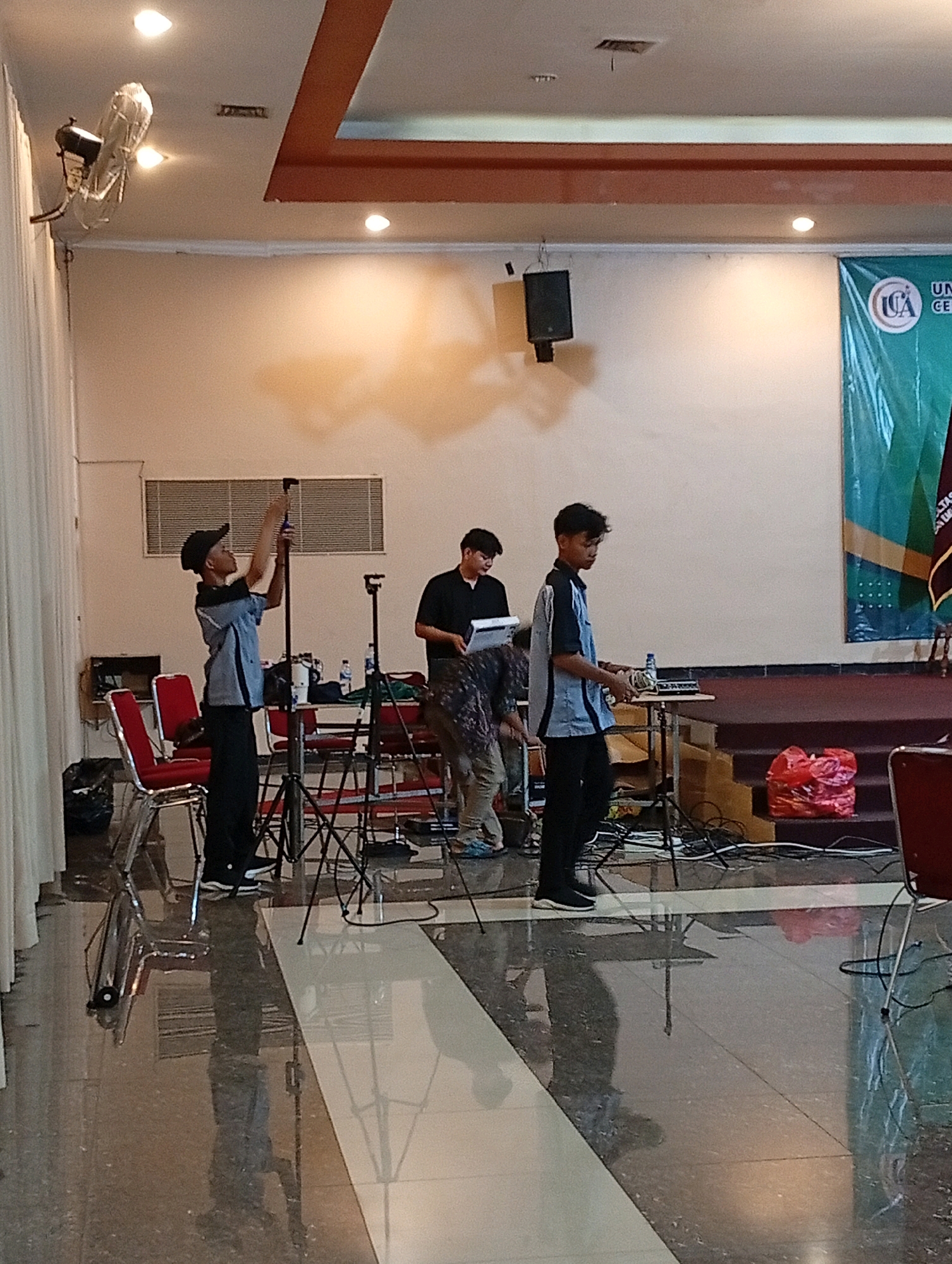 The image shows an indoor event setup with several individuals, likely students or staff, preparing for an activity. In the foreground, a young person wearing a blue and gray shirt, black pants, and a black baseball cap is adjusting a tripod. To their right, another young person in a black shirt and dark pants stands holding a clipboard, looking towards a table with electronic equipment. Further back, a third individual in a black shirt is partially visible behind the person with the clipboard.
The environment appears to be a room with beige walls and a suspended ceiling with recessed lighting. A large banner with a logo that includes "ICA" and text in Indonesian is visible on the right side of the image. Red chairs are arranged on the left, and various equipment, including microphones, speakers, and cables, are set up. The floor is reflective, showing the silhouettes of the people and equipment. The overall scene suggests a preparation stage for a presentation, performance, or meeting, possibly at an educational institution. The presence of the banner with Indonesian text indicates a location in Indonesia, and "Kelapa Dua" is a known area within Indonesia, suggesting this could be a scene from an event taking place in Kelapa Dua.