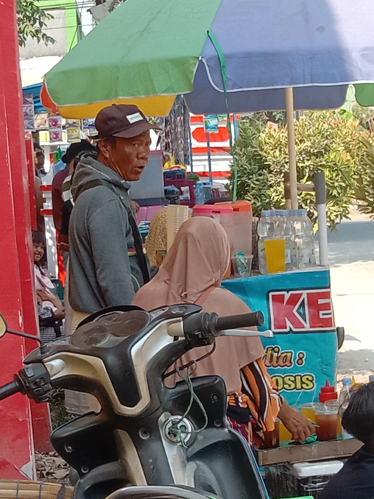 The image captures a street scene in Kunden, Indonesia, focused on a man wearing a dark baseball cap and a grey hooded sweatshirt. He is standing near a market stall or food stand. Behind him, a woman wearing a light brown hijab and a colorful striped shirt is serving drinks. The stall is adorned with colorful umbrellas, a blue and white sign with red lettering that partially reads "KE," and bottles of beverages. A motorcycle is partially visible in the foreground. The background features lush green foliage and other market stalls, suggesting a busy commercial area. The lighting indicates it is daytime, with bright sunlight casting shadows.