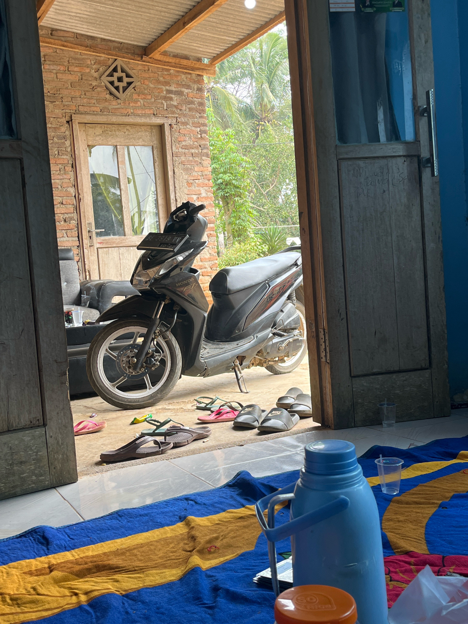 The image depicts a simple, open-air dwelling in Kebumen, Indonesia. The scene is framed by two wooden doorways, one on the left and one on the right, leading to an outdoor area. In the center, a black Honda motorcycle is parked on a concrete floor. The motorcycle has a license plate with the inscription "BE 3919 VV".
Behind the motorcycle, there is a brick wall with a small, decorative vent. A wooden door with glass panes is visible within the brick wall. To the left of the motorcycle, a dark-colored sofa is partially visible.
Scattered on the concrete floor are several pairs of sandals, including pink flip-flops, brown sandals, and black slides. Green and yellow flip-flops are also present.
The foreground of the image shows a colorful blanket or mat with horizontal stripes of blue, yellow, and brown. On the blanket, a large, light blue thermos with two handles is prominently displayed. An orange lid, possibly from a container, is also visible in the foreground. Two clear plastic cups are placed on the blanket.
The background reveals lush green foliage and palm trees, suggesting a tropical environment. The roof of the dwelling is made of corrugated metal supported by wooden beams. The overall impression is a humble, lived-in space in a rural or semi-rural setting within Kebumen. No people are visible in the image.