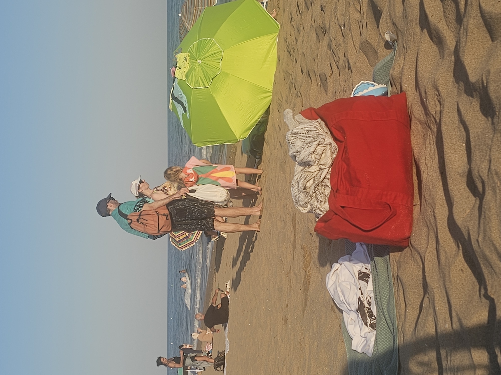 A group of people are enjoying a sunny day at the beach in Castelldefels, Spain. The scene is dominated by a large, vibrant green umbrella providing shade on the sandy shore. Several individuals are visible, some standing and others sitting or lying down.
In the foreground, a man wearing a blue shirt and a dark baseball cap stands with a backpack slung over his shoulder. Next to him, a woman in a patterned dress is visible, as is another person in a pink dress. The sand is marked with shadows, suggesting it is likely late afternoon or early evening.
Further down the beach, people are in the water, with a couple swimming and splashing. The ocean appears calm. Scattered on the sand are various items, including a bright red jacket draped over a towel, a white t-shirt with a graphic print, and other casual beachwear. The overall atmosphere is relaxed and summery, indicative of a typical beach day. The clear sky and warm lighting suggest pleasant weather conditions. No visible text is present in the image.