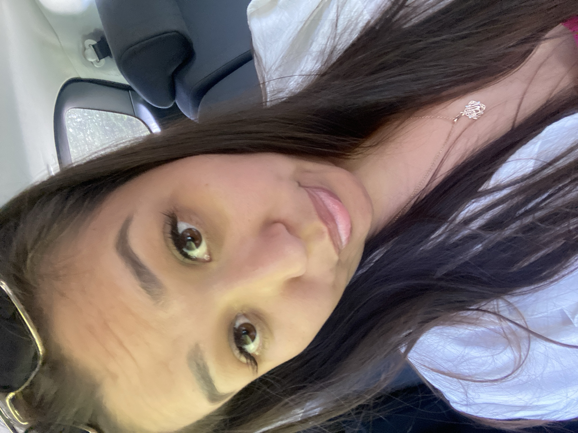 A young woman with long, dark hair and brown eyes is taking a selfie in a car. She is wearing a white top and a necklace with a pendant. She is looking at the camera with a slightly pouting expression. The background shows the interior of a car, including a headrest and a window. It is not possible to determine the location in the city of Zilair, Russia from the image.