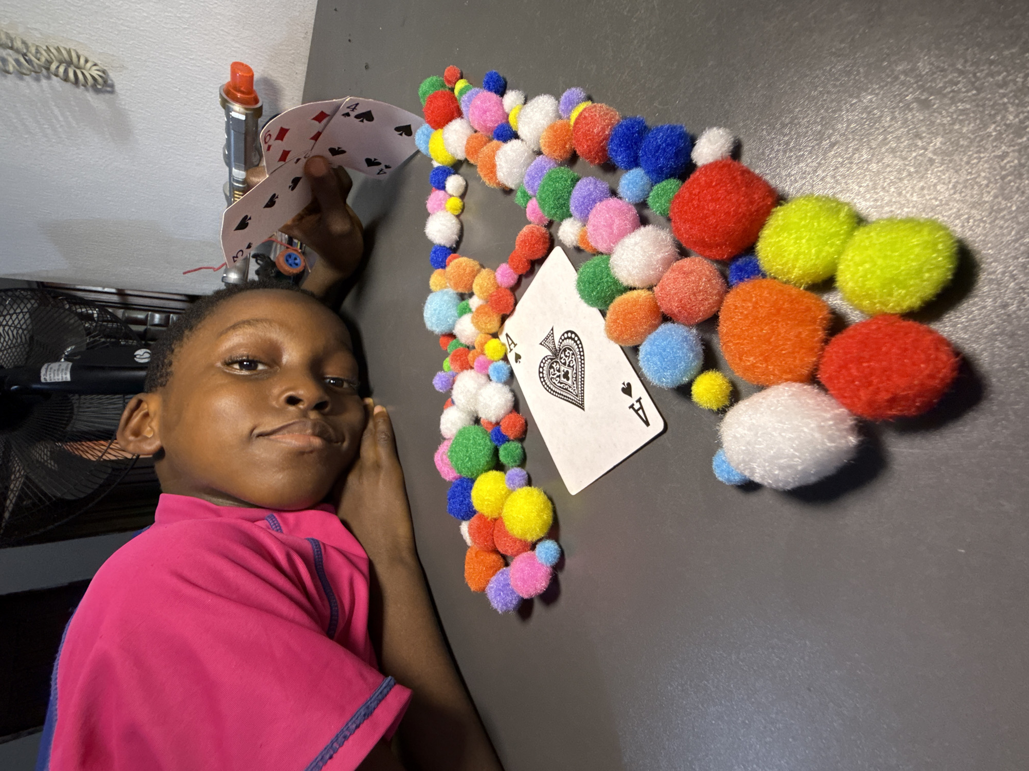 A person lies on a gray surface, their head resting on their hand. They wear a pink shirt. To the person's right, a scattered arrangement of colorful pom-poms surrounds an Ace of Spades playing card. Another hand holds two additional playing cards near a toy or plumbing fixture with a red cap. The background includes parts of a wire mesh object and a light-colored wall. The scene appears to be indoors, and there is no explicit information to link it to Ado, Nigeria.