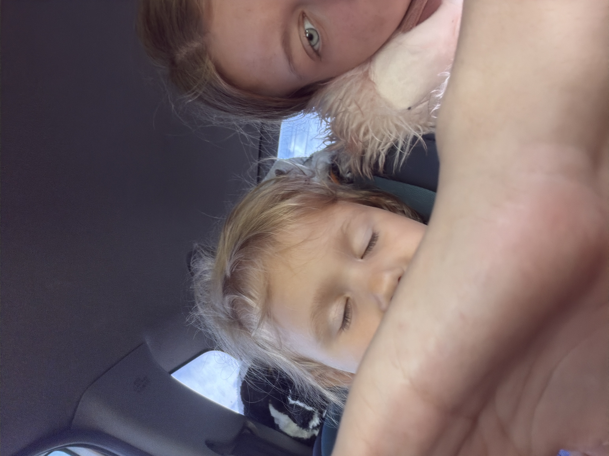 The image shows a close-up view of two individuals in what appears to be the interior of a vehicle. The person in the foreground, likely a child, has blonde hair and their eyes are closed, with their face partially obscured by an arm. To the right, the face of another individual, likely an adult, is visible, with light-colored hair and blue-green eyes. They are wearing a pink, fluffy garment. The background shows the ceiling of the vehicle and a glimpse of the sky through a window. There is no indication of the specific location in the city of Новое Маврино, Russia.