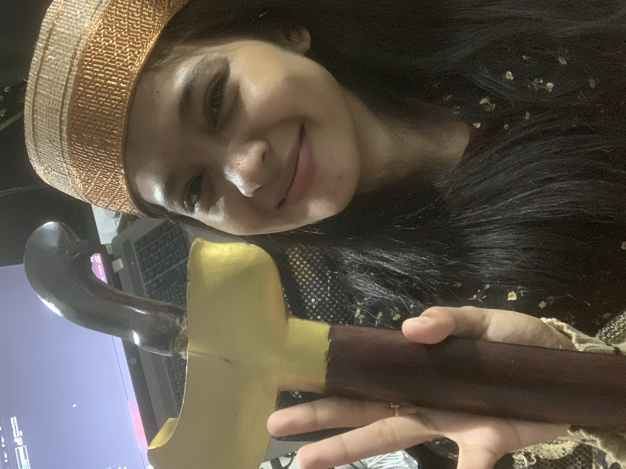 A young woman, presumably in Sungai Meriam, Indonesia, is holding a traditional Indonesian Keris dagger. She is wearing a woven hat and has dark hair. The dagger has a dark brown handle and a golden hilt. A laptop is visible in the background.