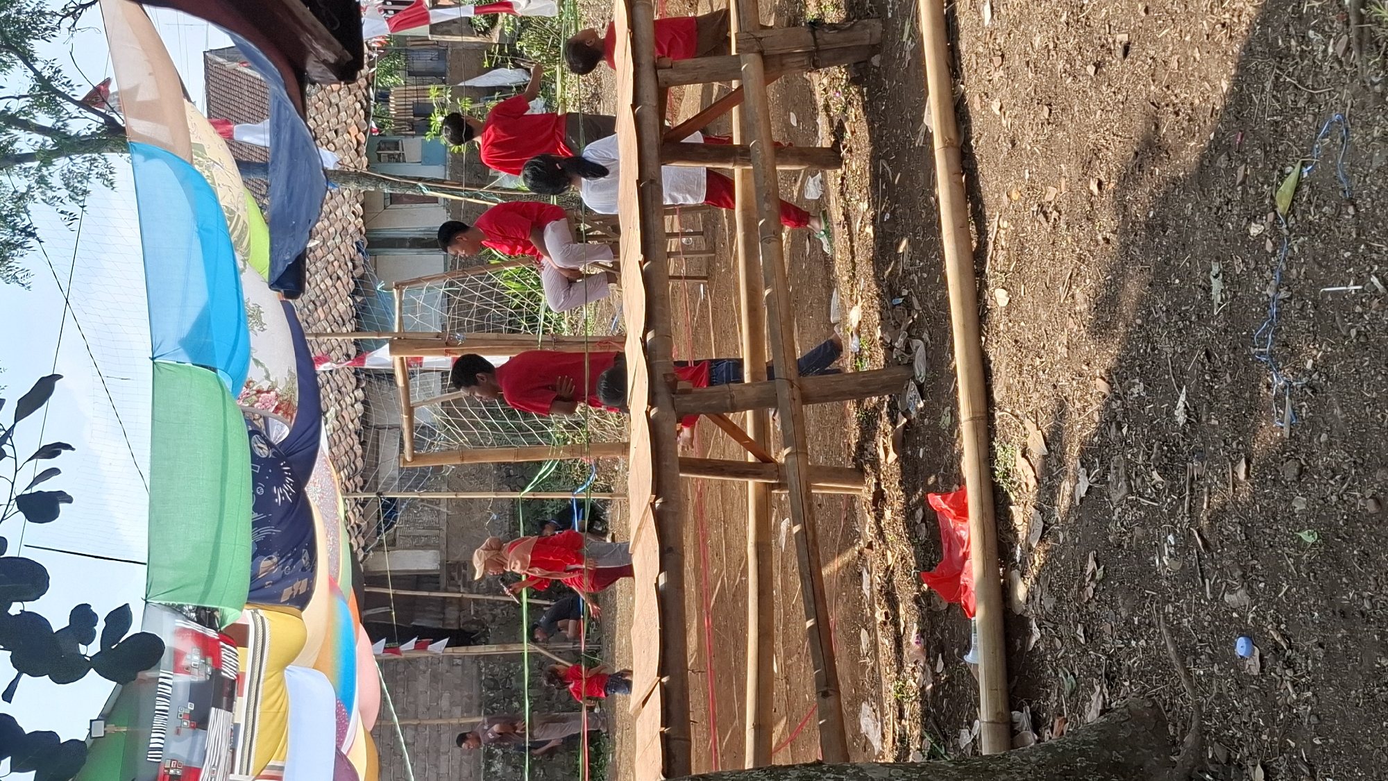 In Cilaku, Indonesia, an outdoor event appears to be underway, possibly a community gathering or festival, with a makeshift stage or seating area constructed from bamboo. Several individuals, primarily young males and females, are present, many dressed in red. Some are standing, while others are seated. In the background, there's a building with a tiled roof and a soccer goal, suggesting a field or open area. Colorful fabrics are draped overhead, creating a decorative canopy. The ground is dirt and littered with some debris, including a red plastic bag and a blue bottle cap. The overall scene depicts a casual, outdoor community event.