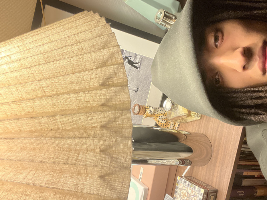 A person wearing a hooded sweatshirt is in the upper right corner, facing the camera. To the left of the person's face is a tan, pleated lampshade. Behind the lampshade are various items on a surface, including a cheetah-print bottle, a shiny silver object, a photograph of people, and a Tiffany & Co. box. Bookcases are visible on the lower right. The orientation of the image is such that the right side is closer to the viewer.