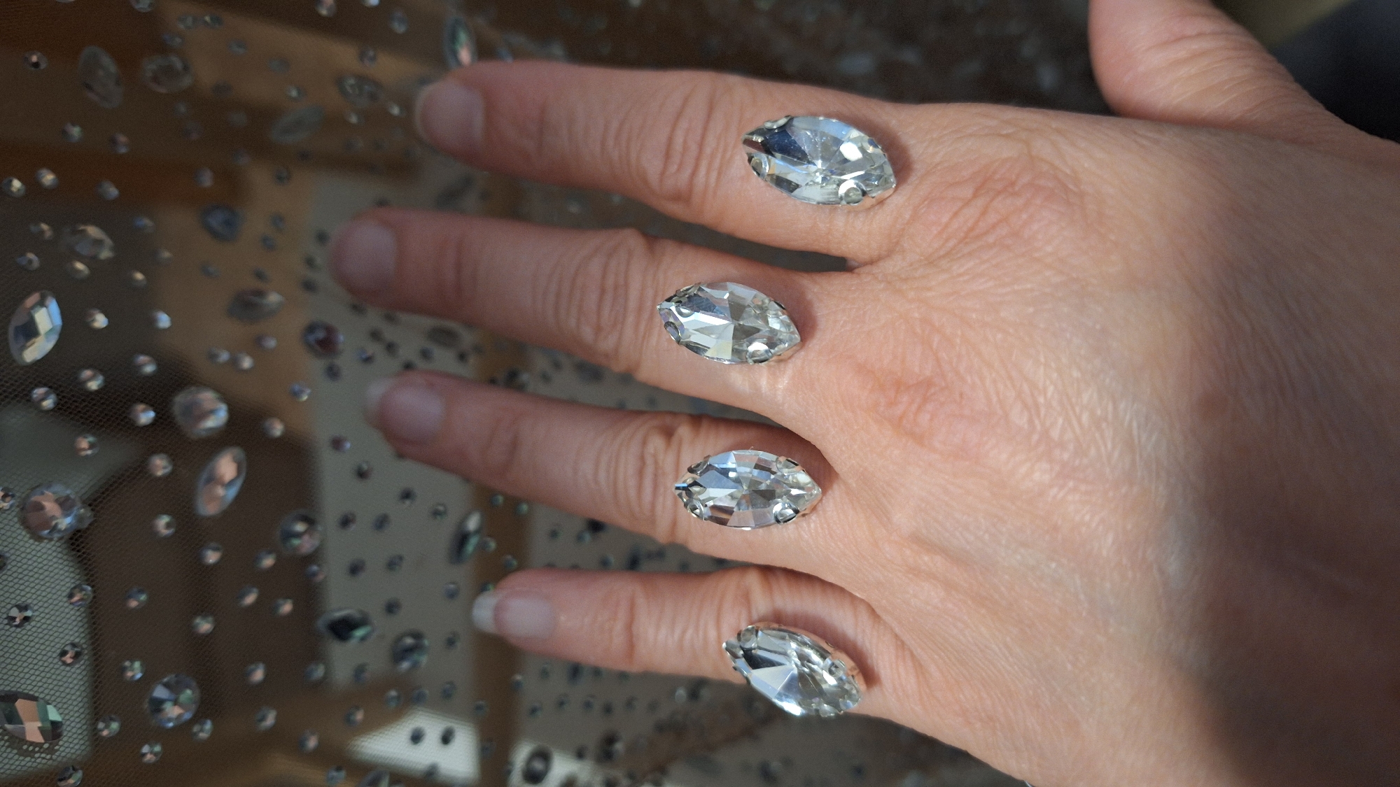 A human hand is positioned against a backdrop of sheer fabric embellished with scattered, clear crystals of varying sizes. The hand features four fingers, each adorned with a single, large, clear, marquise-cut crystal affixed directly to the skin, mimicking the appearance of rings. The fingernails have a natural, light-colored manicure. The hand is posed in a way that suggests interaction with the fabric. The lighting is bright and direct, highlighting the facets of the crystals and the texture of the skin. The background is blurred but seems to be a decorative textile with more crystals on it. There are no explicit indications of the location being Blagoevgrad, Bulgaria.