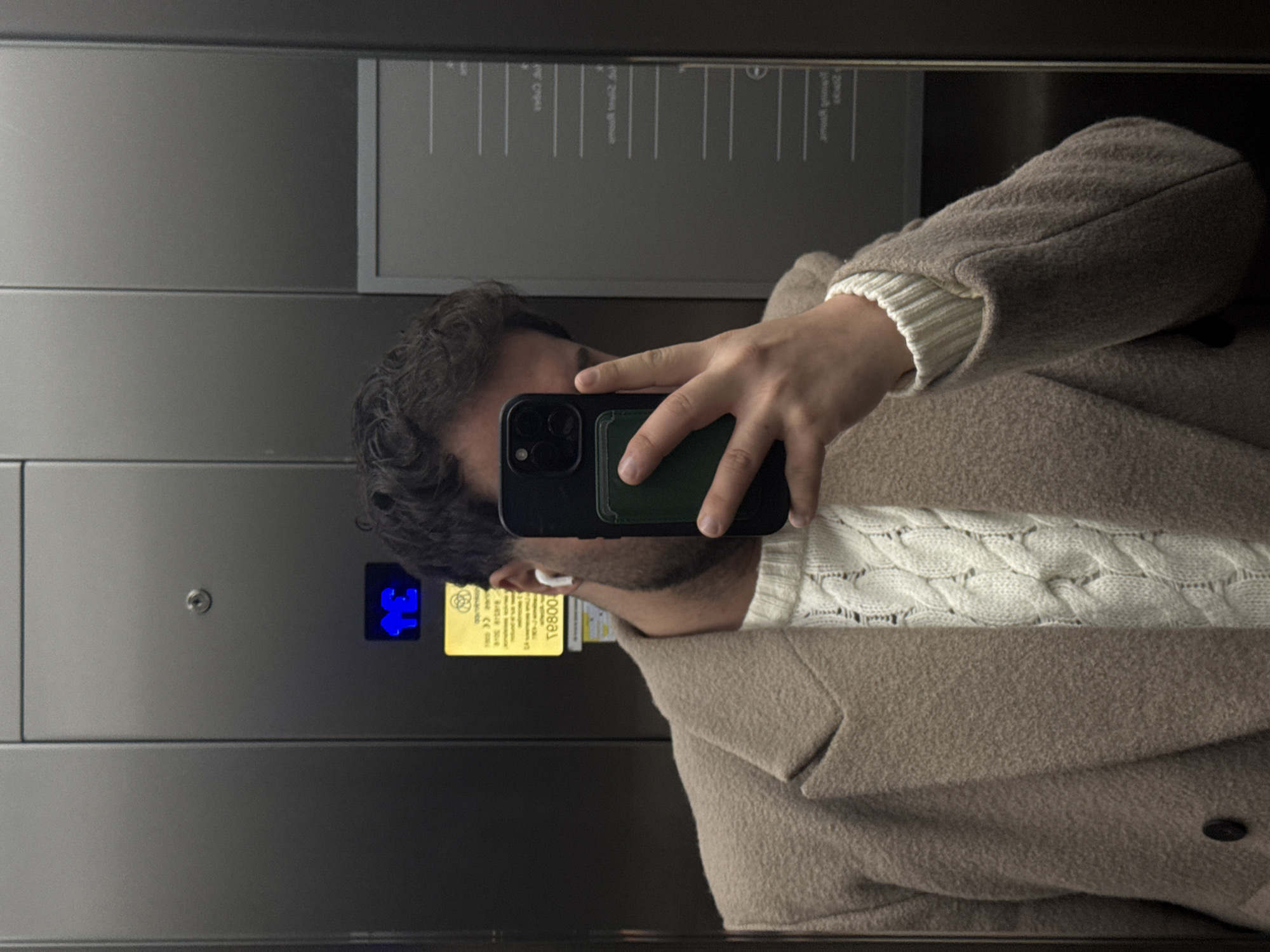A person takes a selfie in an elevator. The person is wearing a white cable-knit sweater and a tan overcoat. They are holding a black smartphone with a green card case. The elevator has grey metal walls and a panel with a blue illuminated figure of a person and a yellow information sign. Another panel shows text possibly in French and German. The location is likely in Esch-sur-Alzette, Luxembourg.