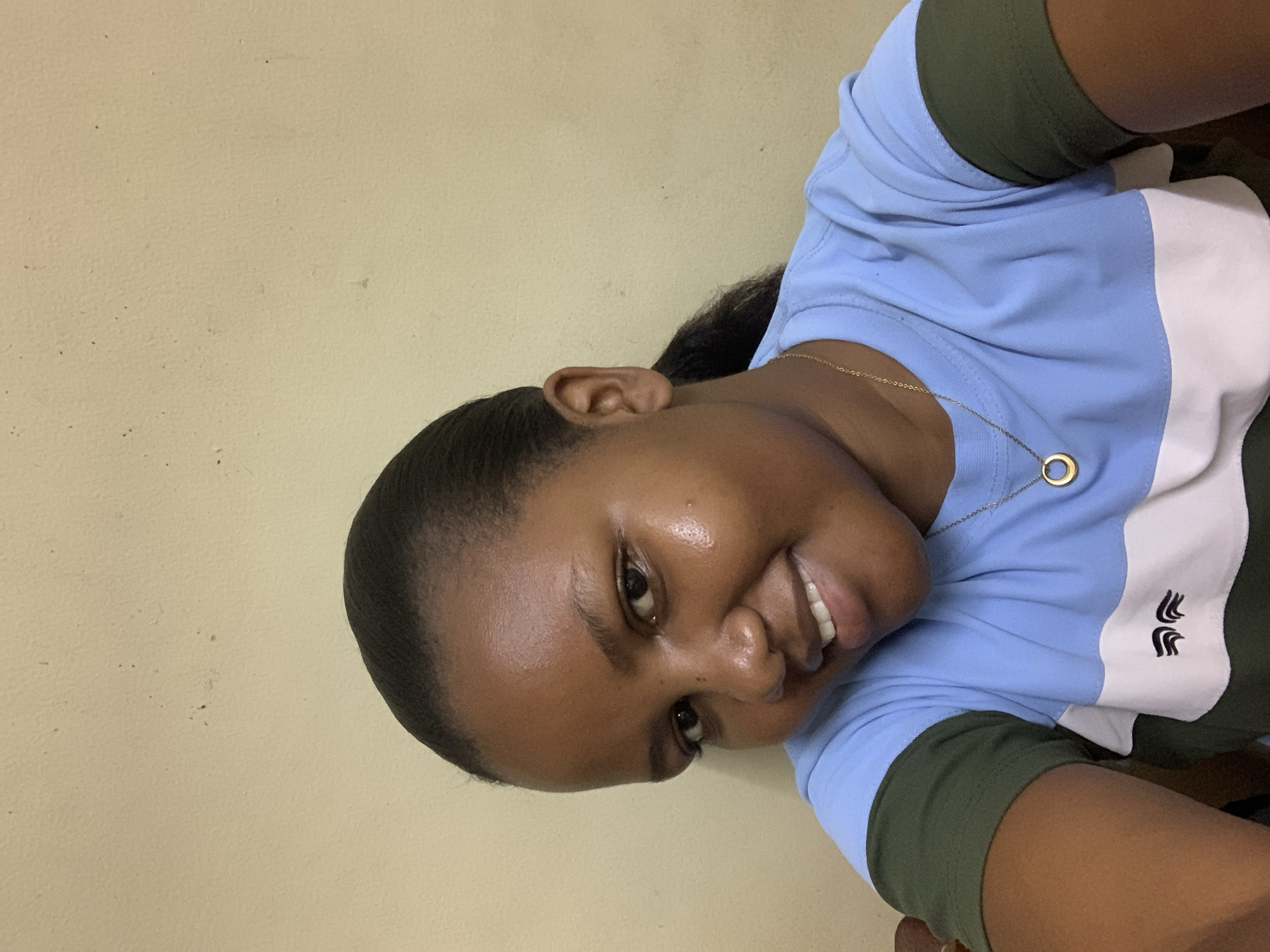 A young person, appearing to be female, is captured in a selfie-style portrait, with the image rotated 90 degrees clockwise. She has dark skin and her dark hair is pulled back. She is smiling broadly, revealing her teeth, and her eyes are looking towards the camera. Her expression conveys happiness and friendliness.
She is wearing a color-block polo shirt that features horizontal stripes of light blue, white, and olive green. A simple, gold-colored necklace with a small, circular pendant rests on her collarbone over the blue section of her shirt. On the white stripe of her shirt, near her right shoulder, there is a small black embroidered logo consisting of three stylized, upward-curving elements, possibly resembling abstract feathers or wings.
The background is a plain, light-colored wall, likely cream or very light yellow, with some minor imperfections visible. The lighting appears even, suggesting an indoor setting. There are no other objects or people visible in the frame. The scene is set in what is understood to be Dar es-Salaam, Tanzania.