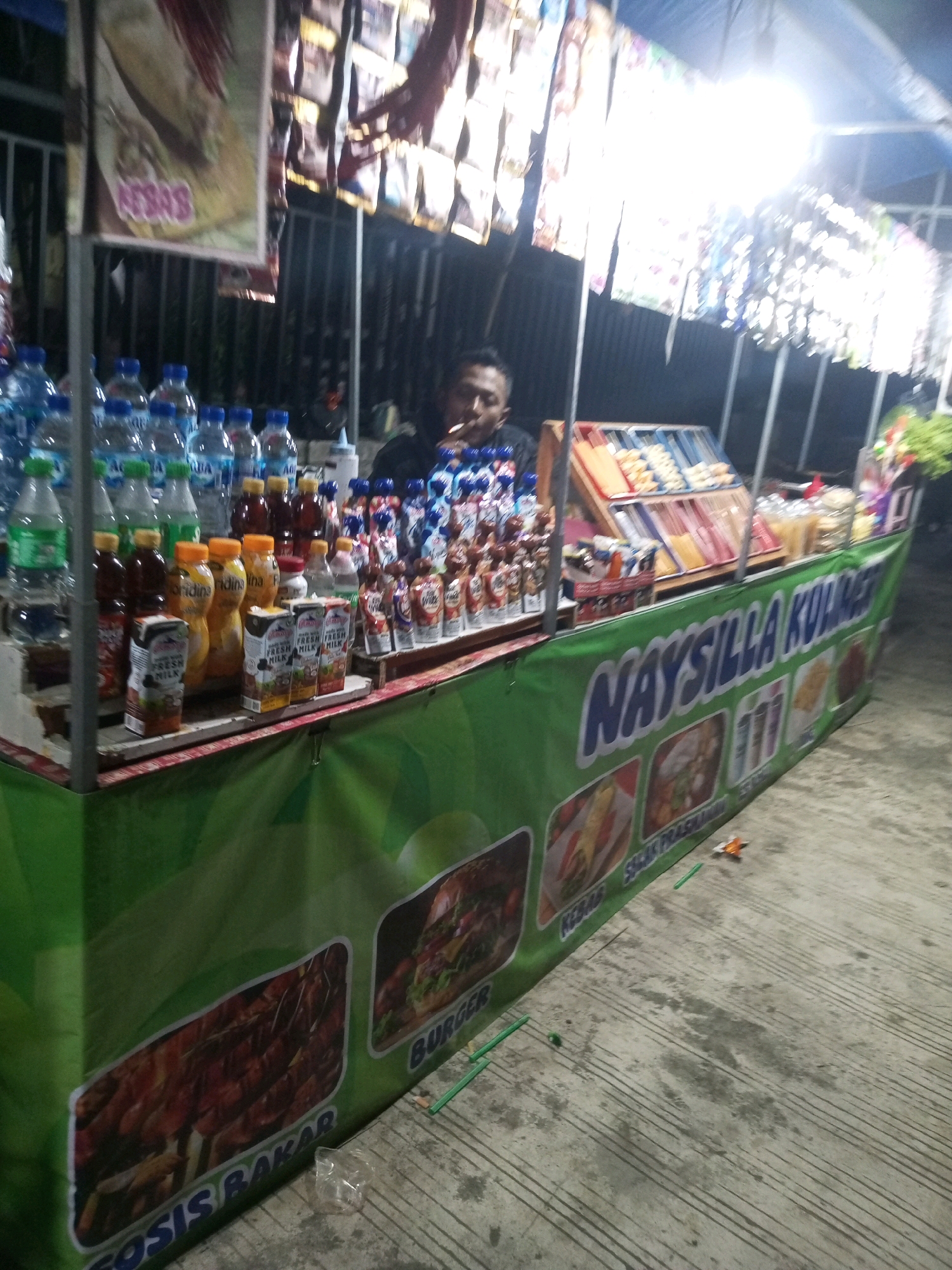 The image depicts a street food stall in Wonosari, Indonesia, at night. The stall is illuminated by artificial lights, creating a bright, slightly overexposed scene. A vendor, a man with dark hair and a t-shirt, is partially visible behind the counter, smoking a cigarette. The stall displays a variety of products, including bottled drinks, juice boxes, and packaged snacks. The front of the stall features a large green banner with white lettering that reads "NAYSILLA KUHAAJ", and various food images are displayed along the bottom of the banner, including grilled sausages, burgers, and what appear to be noodles. The ground is concrete and appears wet in places. The overall impression is that of a busy, albeit dimly lit, night market or roadside food vendor.