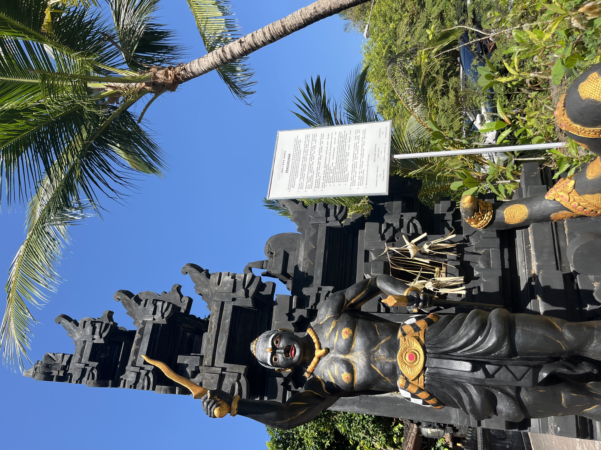An eye-level, medium shot captures a black, imposing statue of a warrior in traditional Balinese attire, standing in front of a dark, ornate temple structure under a clear blue sky. The statue is detailed with gold accents, including a chest adornment, a belt with a red gem, and armbands. It holds a golden dagger in its raised right hand and another in its left. The warrior's face is painted with white markings and red lips. In the foreground, two golden serpent-like figures with yellow scales and red eyes emerge from the base of the statue. To the right of the statue, a white informational sign with Indonesian text is mounted on a metal pole, surrounded by lush green foliage and a palm tree. The background reveals more greenery and the roof of a vehicle. The overall scene suggests a cultural or religious site. The location is identified as Kuta Selatan, Indonesia.