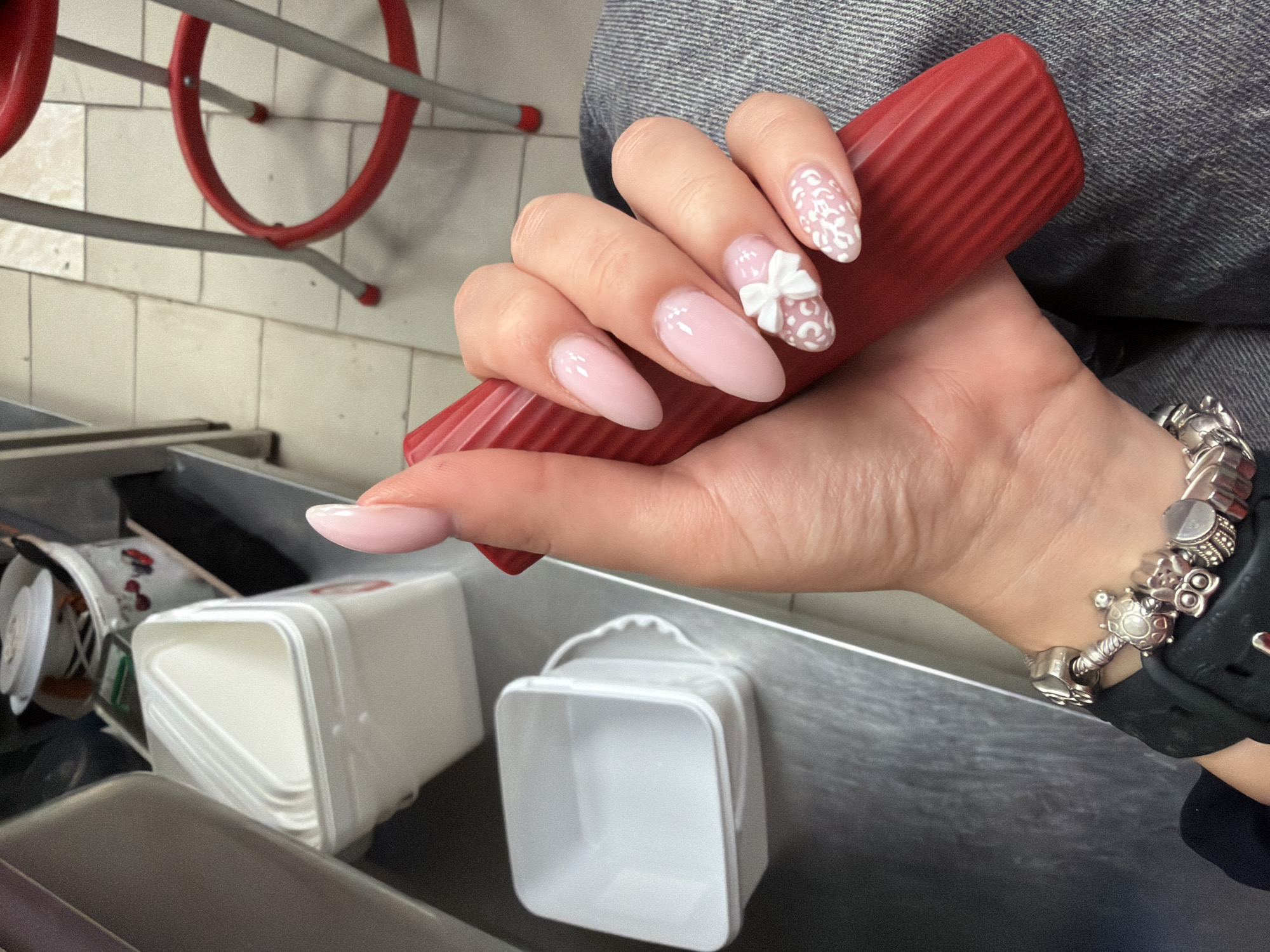 The media file is a close-up shot of a person's hand with a light pink manicure, holding a ridged red object. The nails are long and almond-shaped. Two of the nails feature intricate white lace-like designs, while one has a small, white 3D bow accent. The hand also wears a silver charm bracelet with various charms, including a turtle.
In the background, out of focus, are elements that suggest an indoor, possibly commercial or industrial setting. There are metal shelves or countertops, and stacked white containers. Some red metal structures, possibly part of a rack or railing, are visible in the upper left corner. The lighting suggests it is daytime, and the overall tone is neutral. The location context provided is Plovdiv, Bulgaria. No visible text is present in the image.