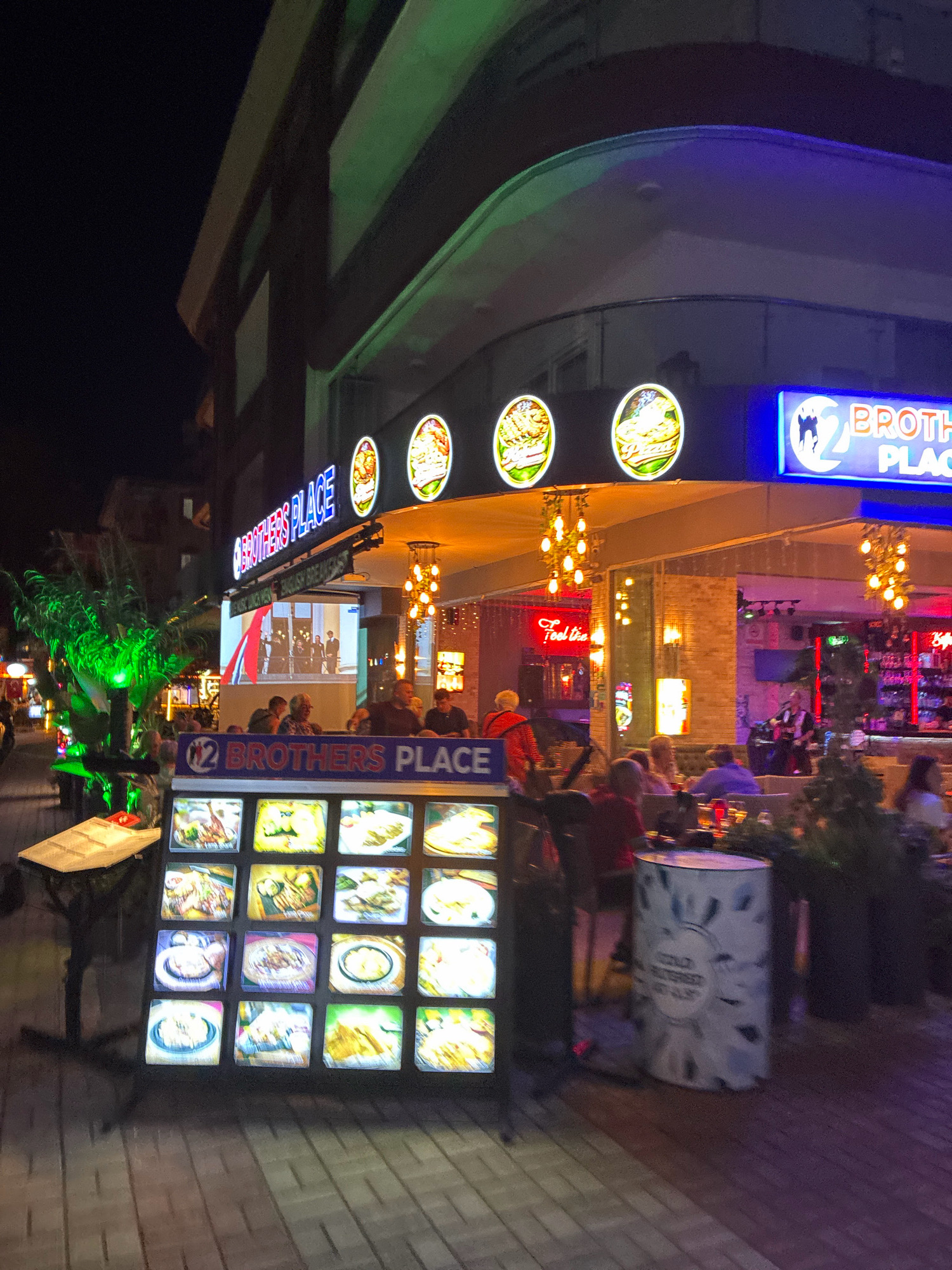 The image shows the exterior of a restaurant at night, likely in Alanya, Turkey, indicated by the text on the building's signage and a displayed menu. The restaurant is named "BROTHERS PLACE" and features illuminated signs with food imagery, including pizzas and what appear to be burgers. A large menu board displays various food items in illuminated squares.
Several patrons are seated at tables outside the restaurant, enjoying their meals and drinks. Some individuals are visible inside the establishment, which appears to be a bar or lounge area, with bottles and glasses displayed. A live musician is performing on a small stage inside. The overall atmosphere is lively, with the restaurant illuminated by bright, colorful lights and decorative lamps hanging from the awning. The street pavement is visible in the foreground, and a trash receptacle with a patterned design is situated near the outdoor seating area.