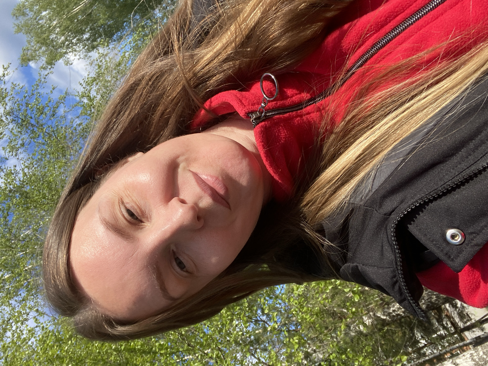 A woman with light skin and brown hair is pictured from a low angle. She is wearing a red fleece jacket under a black jacket. The zipper of the red jacket is visible. The background shows trees with green leaves and a blue sky with white clouds. The photograph appears to have been taken outdoors in daylight. There is no visible evidence to confirm or deny the location is in Noviy, Russia.