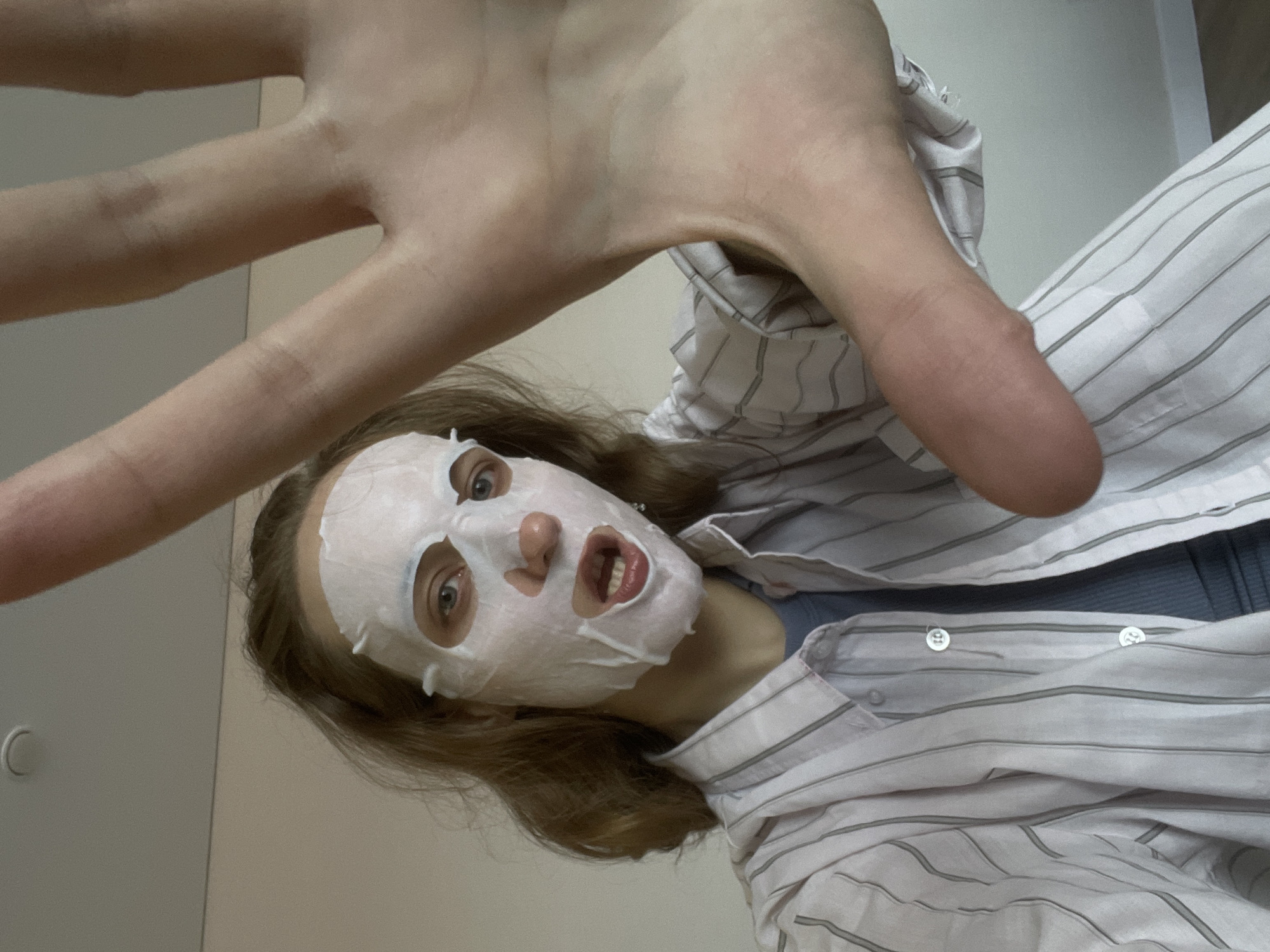A Caucasian woman, appearing to be in her late teens to early twenties, is wearing a white facial mask and a striped button-down shirt. Her right hand is outstretched towards the camera. The background is a pale wall with a white door. The photo appears to be taken indoors. No city information.