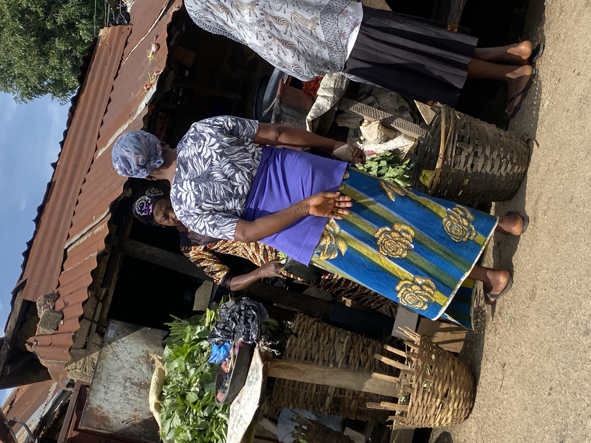 The image depicts an outdoor market scene, likely in the city of Kabong, Nigeria, given the context. The focus is on three individuals, presumably women, interacting at a stall with a corrugated metal roof. In the foreground, a woman wearing a blue and yellow patterned skirt and a white and black leaf-patterned shirt is seen from behind, with her hands clasped behind her back. To her left, a woven basket is visible. Further back and to the left, a woman in a brown and gold patterned outfit appears to be interacting with the woman in the foreground, or tending to the goods. She is wearing a headscarf. To the right of the woman in the foreground, another individual is partially visible, wearing a light-colored patterned top and a black skirt. The stall itself features an abundance of fresh green leafy vegetables displayed in a shallow bowl and within woven baskets. Various other items, including what appear to be plastic bags and possibly other produce, are also present at the stall. The ground is a dusty, paved surface. The overall impression is of a bustling marketplace with a focus on fresh produce.