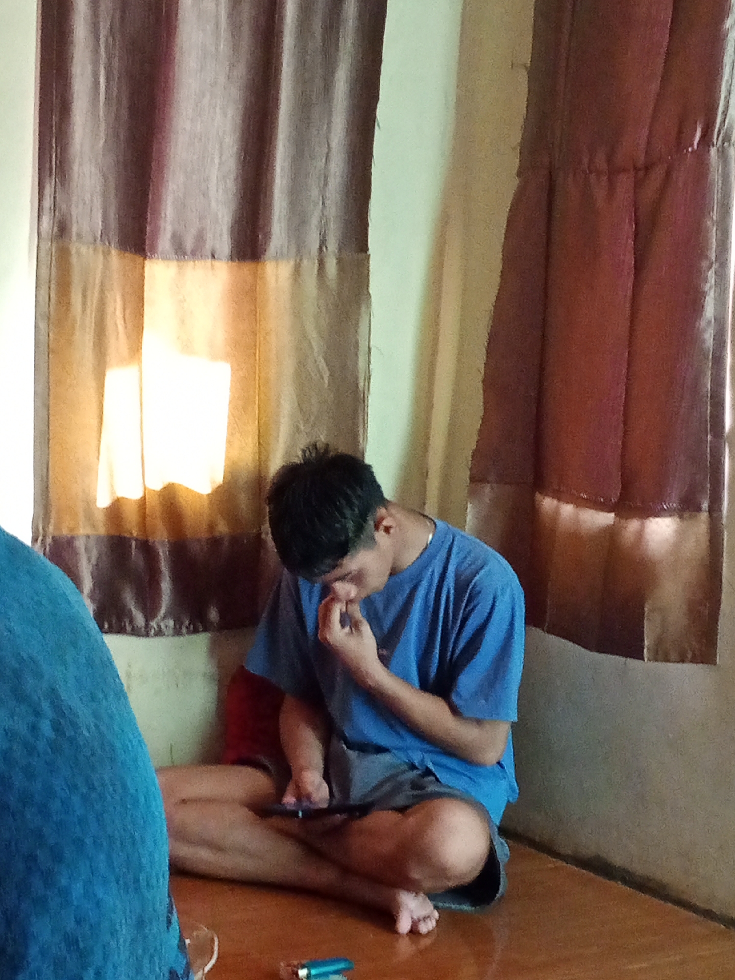 A young man is sitting cross-legged on the floor in front of a window covered by a light brown and dark maroon curtain. He is wearing a blue t-shirt and grey shorts and is looking down at a black object, possibly a phone, in his hands. His right hand is touching his face, near his nose. To the left of the frame, a portion of a large, blue object, likely a piece of furniture or clothing, is visible. The floor is a reddish-brown wooden surface. A small silver and blue object, resembling a lighter, is on the floor to the right of the man. The lighting suggests it is daytime, with light filtering through the window. The scene appears to be indoors, in a residential setting. The location is Subang, Indonesia.