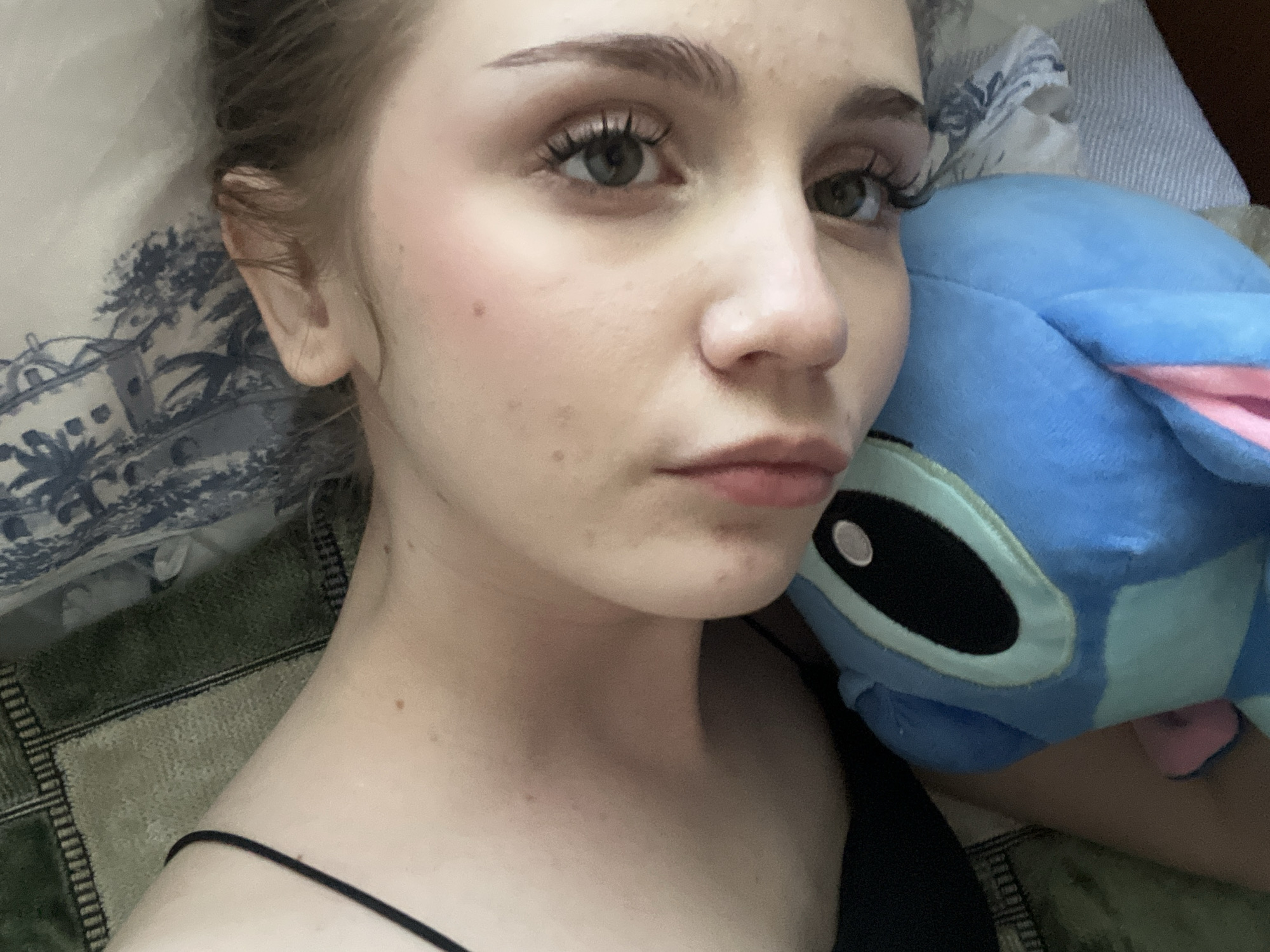 A close-up, overhead shot shows a young woman lying down, with her head resting on a pillow and a stuffed toy. The woman has light skin, green eyes, and her hair is pulled back. She is wearing light makeup, including blush and mascara. She is looking directly at the camera.
The stuffed toy is blue and appears to be a character from a cartoon, possibly Stitch from Lilo & Stitch, with large black eyes and pink accents. The background includes a patterned pillow and a textured blanket. The lighting suggests it is daytime indoors. The woman's expression is neutral, but she appears relaxed.
The context is Ashkadar, Russia. No text is visible in the image.