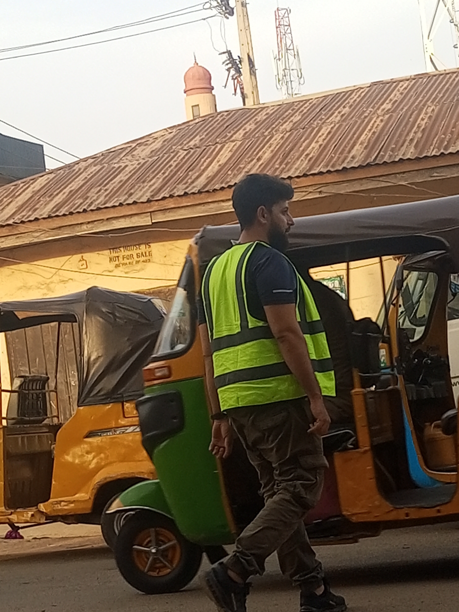 The image shows a man in a lime green safety vest walking past a yellow and green auto rickshaw, a common form of transportation in Maiburiji, Nigeria. The building behind him has a corrugated iron roof and a sign reading "THIS HOUSE IS NOT FOR SALE." In the background, a minaret and telecommunications tower are visible. The setting appears to be a busy street.