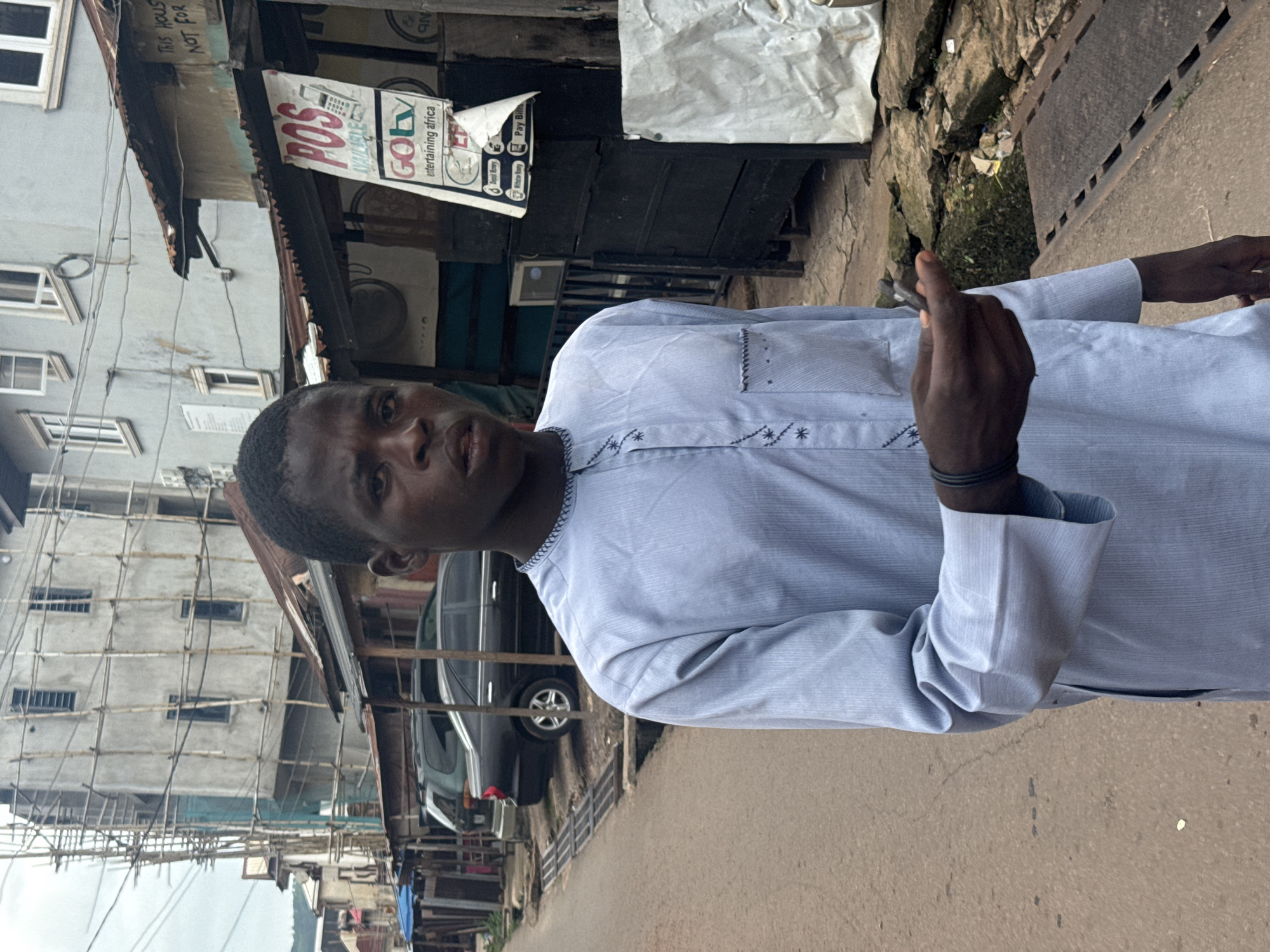 A young Black male, wearing a light blue dashiki with decorative stitching and an embroidered pocket, stands on a dirt road in an urban environment. He is looking directly at the camera with a neutral expression and holding a small, dark object in his right hand. Behind him, a narrow street is lined with various structures. To his left, a multi-story building is under construction, covered in scaffolding. Further down the street, several parked cars are visible. To his right, a row of single-story shops with corrugated metal roofs is present, including one with a prominent "POS AVAILABLE" sign and "GOTV entertaining africa" branding. The ground is uneven, with visible drains and debris. The overall scene suggests a bustling, developing area within the city of Enugu, Nigeria.