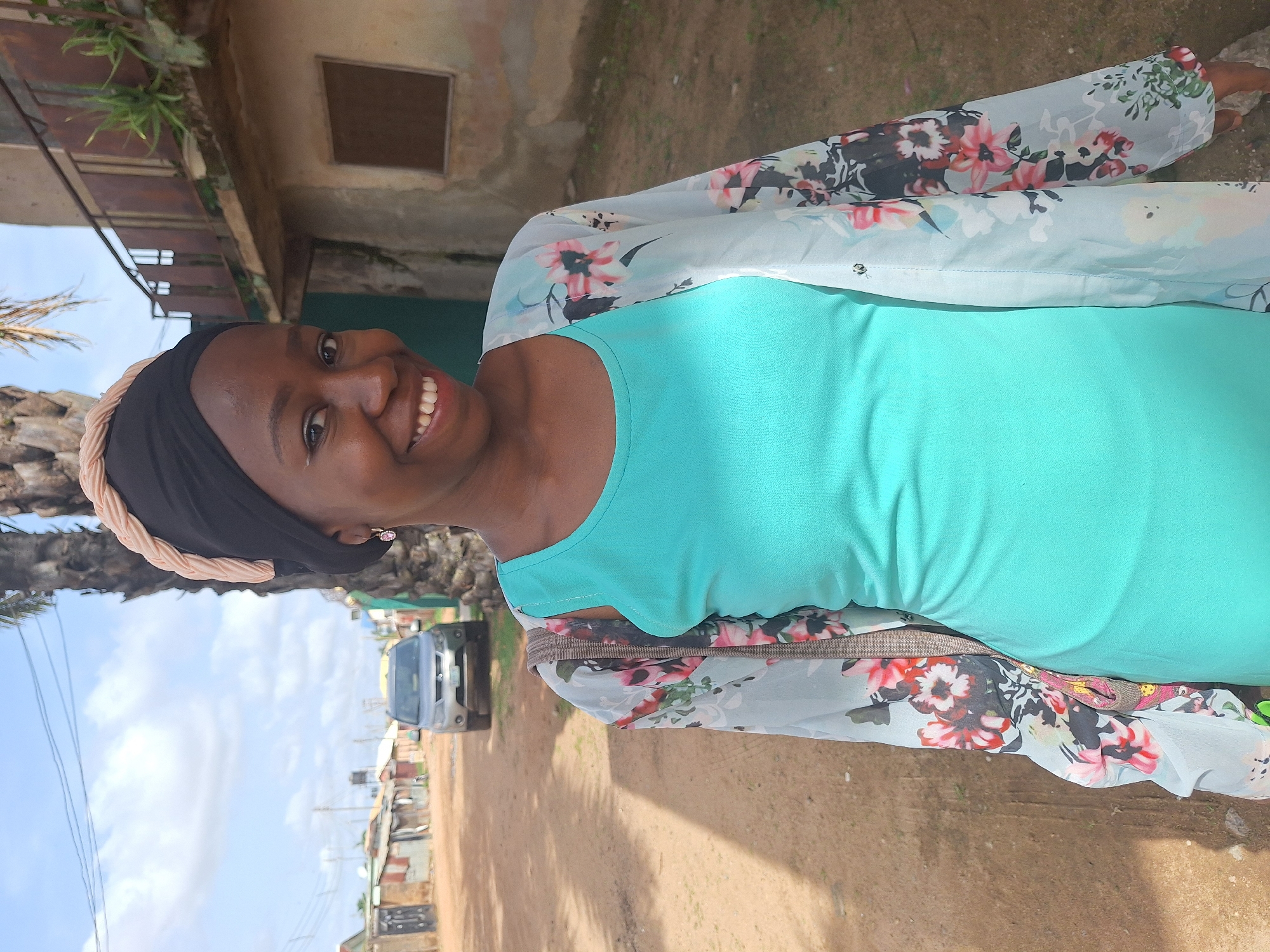 A smiling Black woman stands outdoors on a dirt road in Sabon Tasha, Nigeria. She wears a bright turquoise dress, a sheer floral kimono, and a black headscarf with a peach-colored braided border. Buildings line the road, a grey van is parked in the background, and power lines are visible in the sky. The sky is blue with light clouds.