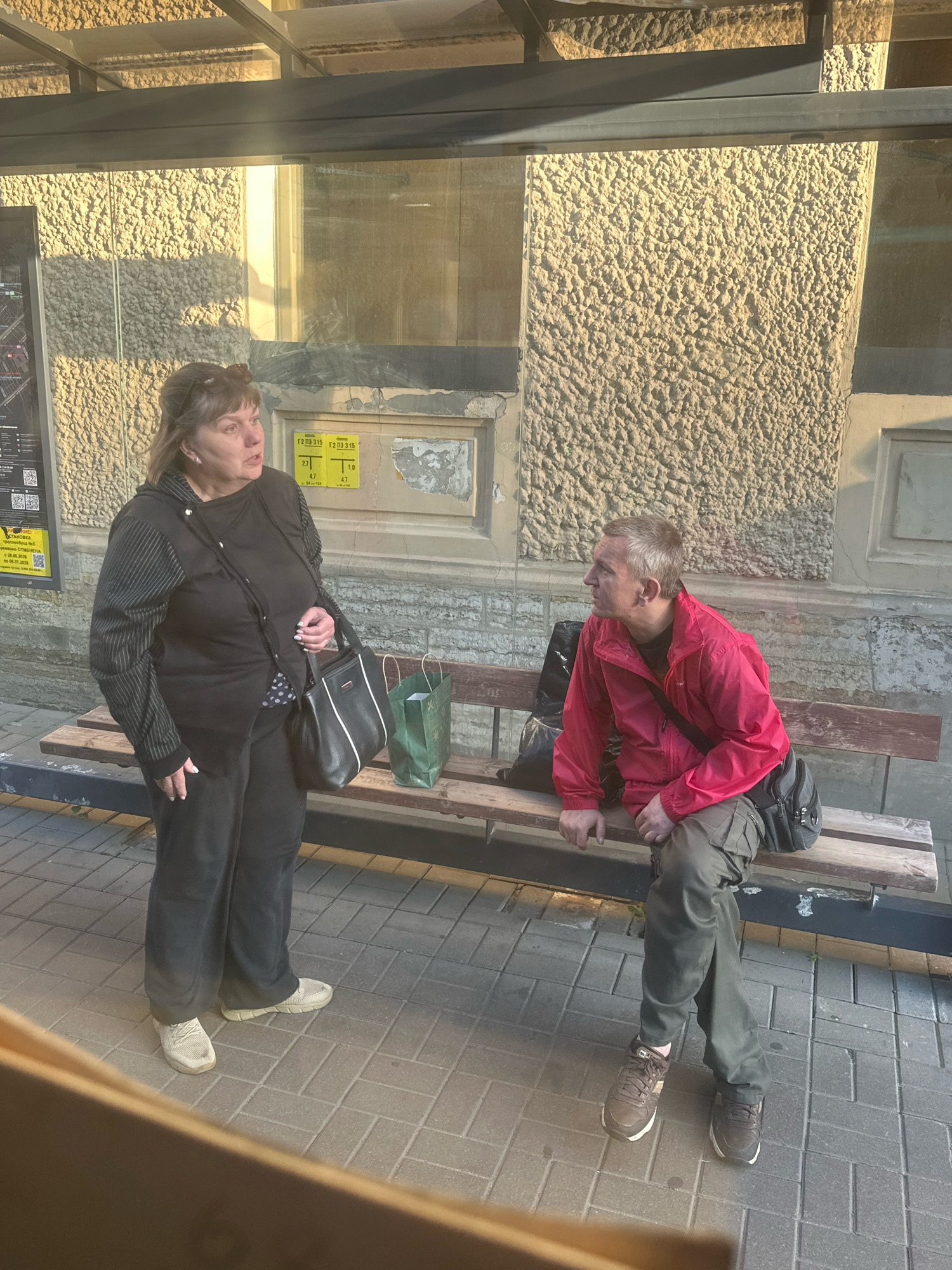 An outdoor scene at what appears to be a bus stop in Saint Petersburg, Russia, features two individuals on a wooden bench. To the left, a woman with blonde hair pulled back is standing. She is wearing a black zip-up jacket with striped sleeves over a black top, and dark pants. She carries a black handbag with white trim. To the right, a man with short, graying hair is seated on the bench. He is wearing a bright red jacket over a dark shirt and khaki-colored cargo pants. A dark bag is slung over his shoulder. A green shopping bag rests on the bench between the two people, and a black bag is behind the man. The background consists of a textured, beige wall and a glass-covered shelter overhead. The ground is paved with light-colored brickwork. The lighting suggests it is daytime, with shadows cast by the shelter and the woman.